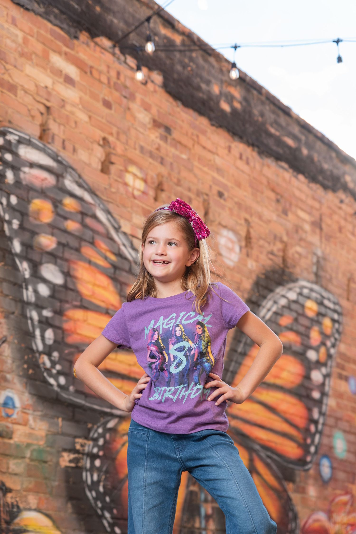 Girl in purple birthday shirt poses confidently in front of colorful butterfly mural on brick wall.