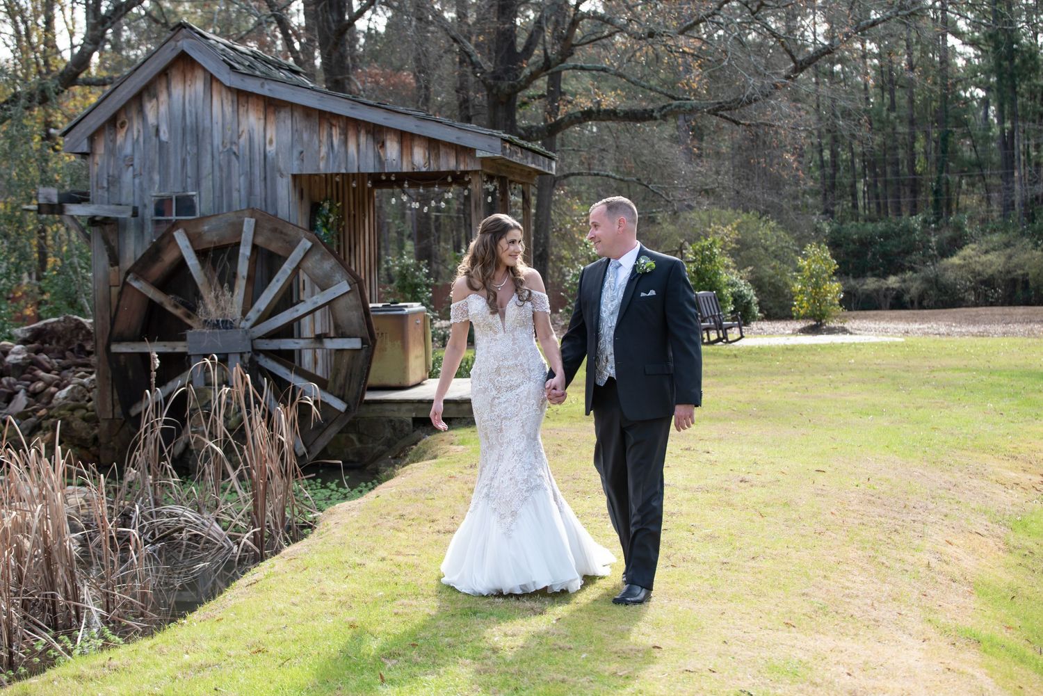 Couple walks together near rustic water mill building during wedding photo session.
