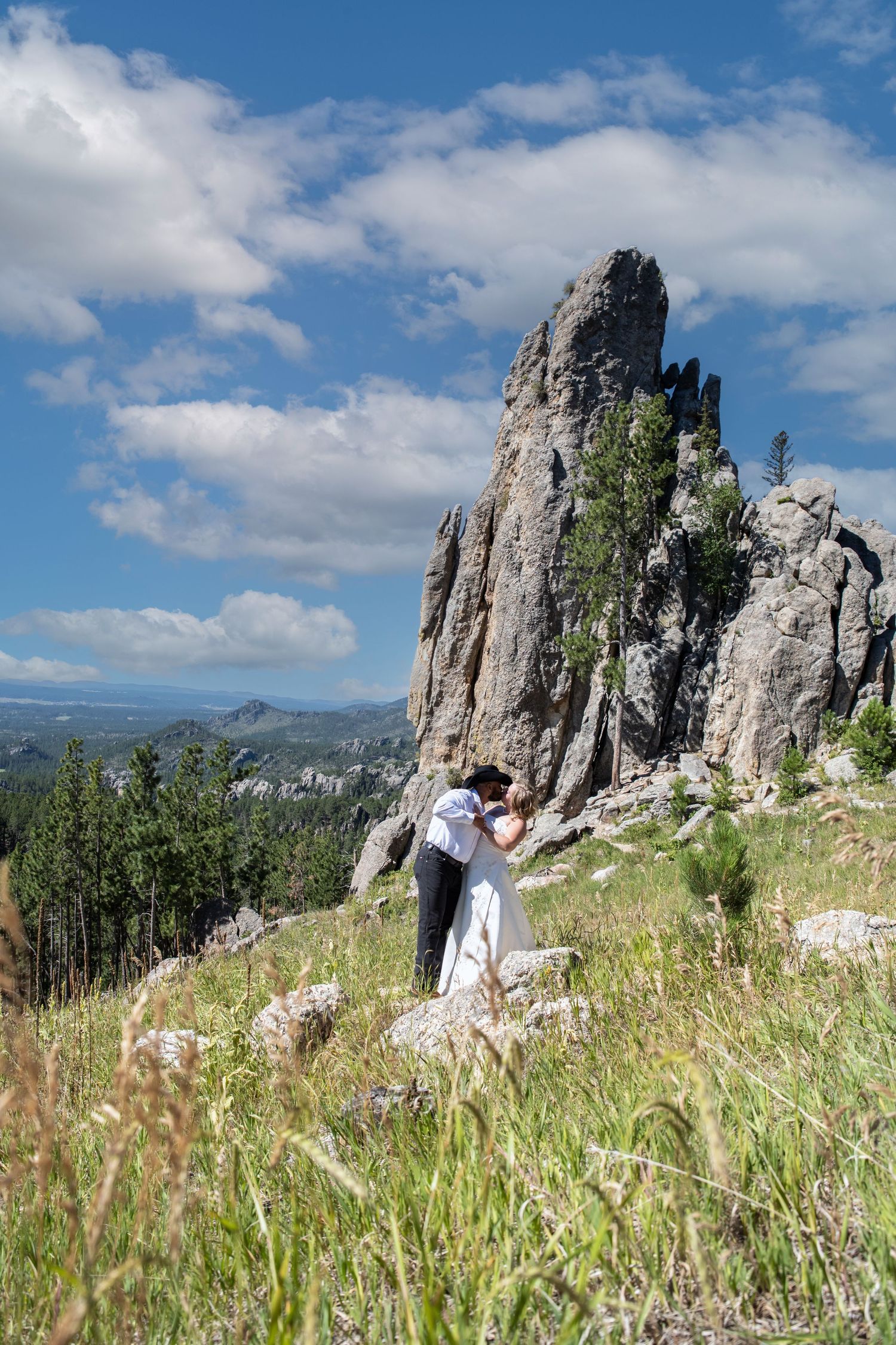 Wedding couple embraces on mountain summit with dramatic rock formation and scenic vista.