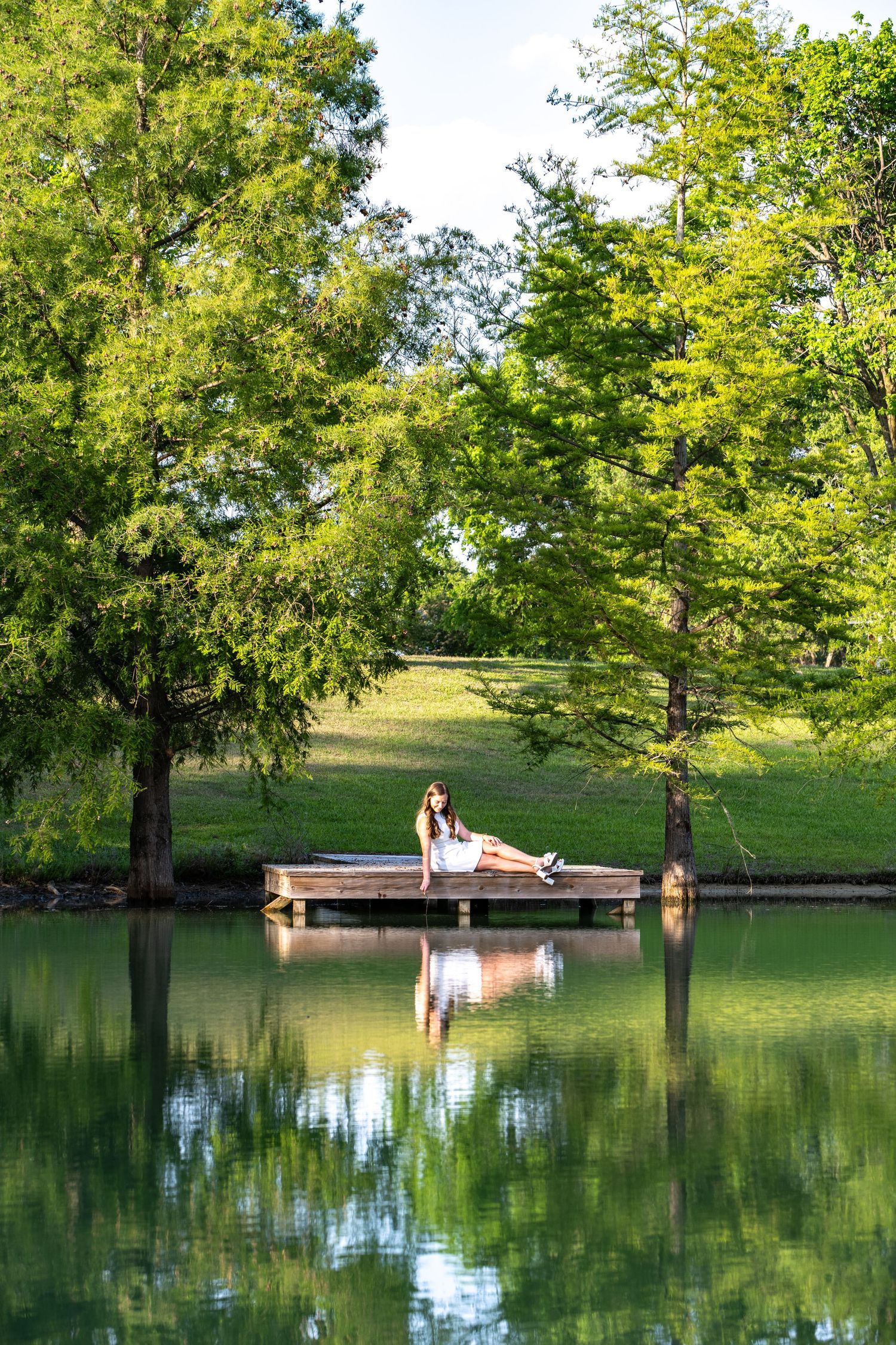 Woman sitting on a wooden dock by a calm reflective pond surrounded by lush green trees in a serene park setting.