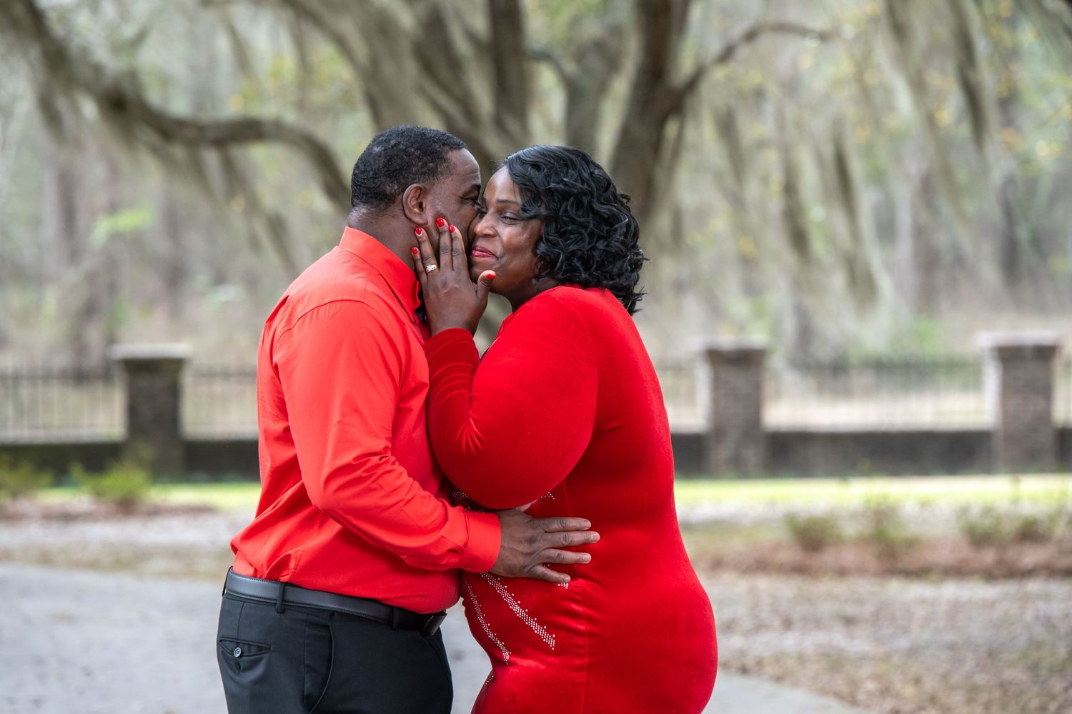 A romantic couple dressed in matching red outfits embrace tenderly in a misty park with Spanish moss hanging overhead.