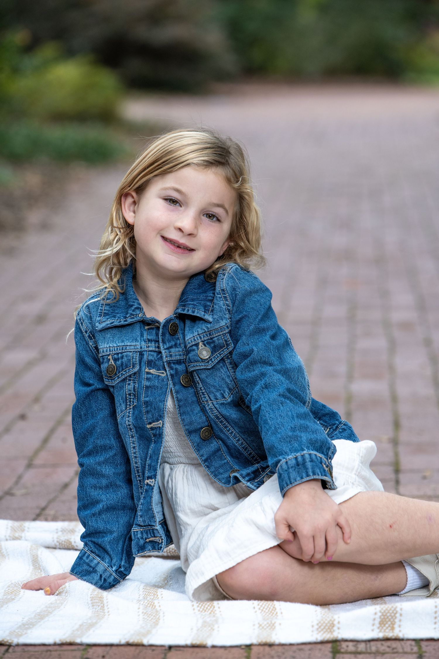 Young girl in denim jacket and white dress sitting on blanket outdoors, smiling at camera.