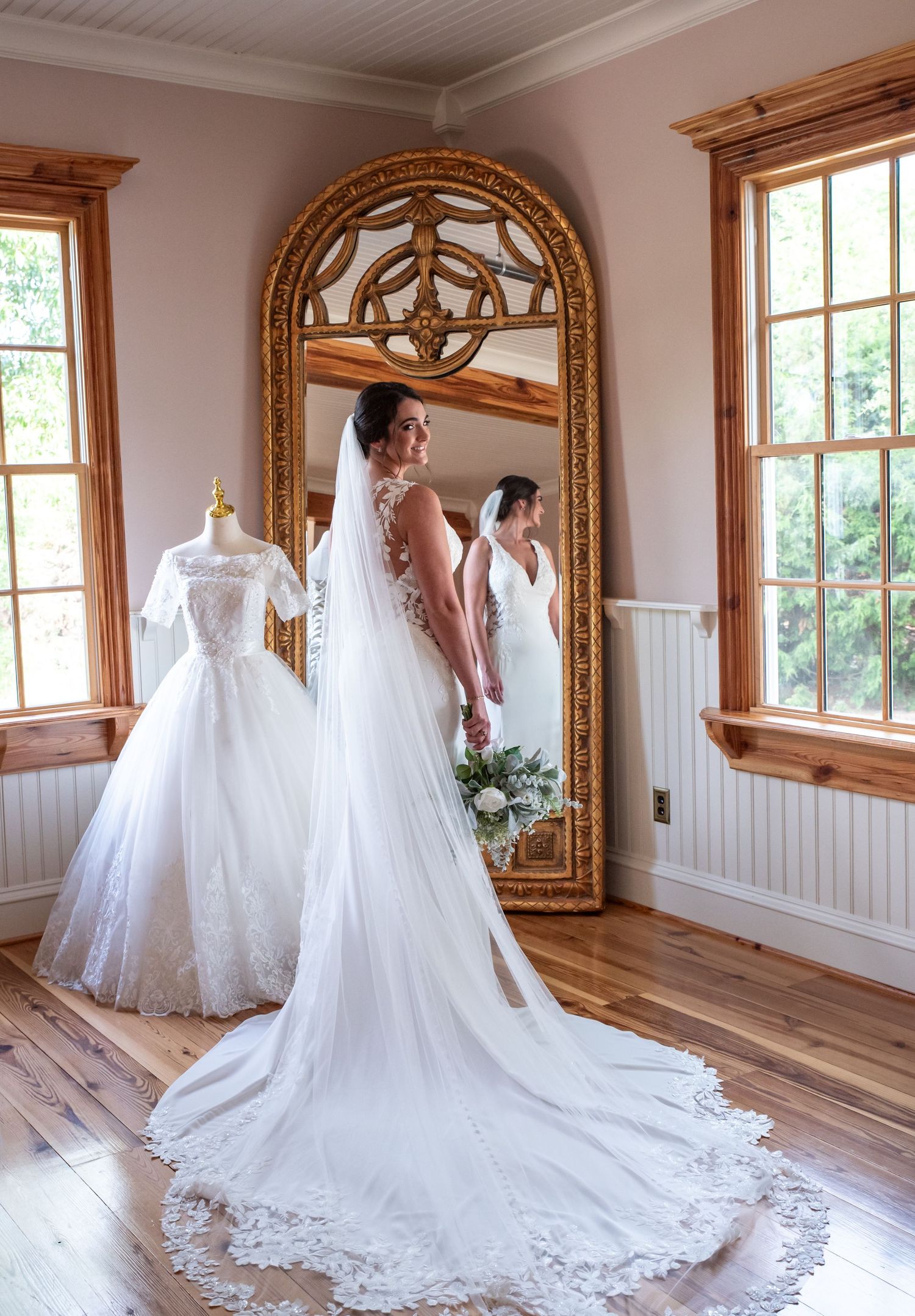 Bride in white gown with long train stands before ornate gold mirror, with vintage dress displayed on mannequin nearby.