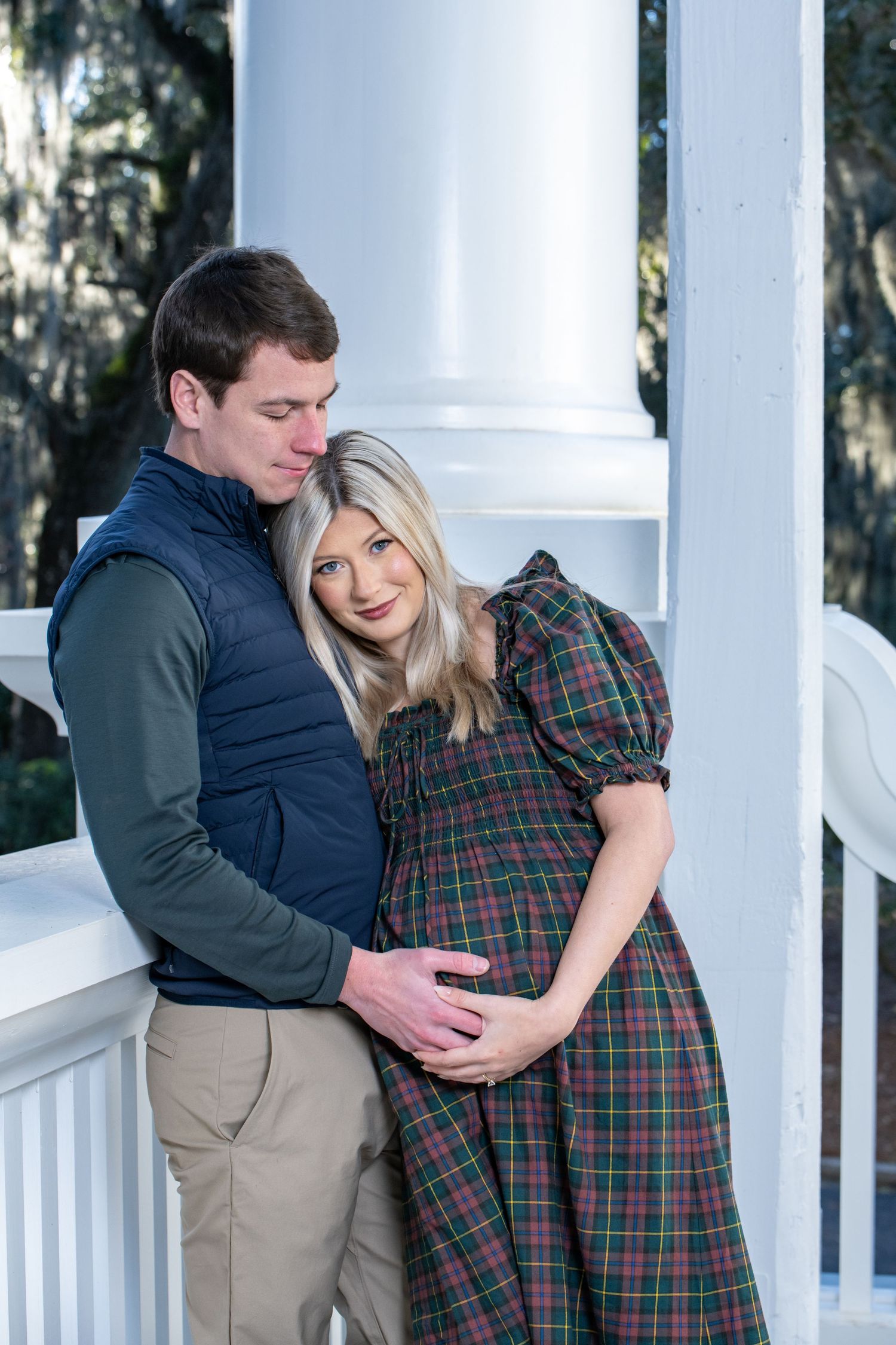 Couple sharing a tender moment on a white porch, woman in plaid dress cradling baby bump, man in navy vest embracing her.