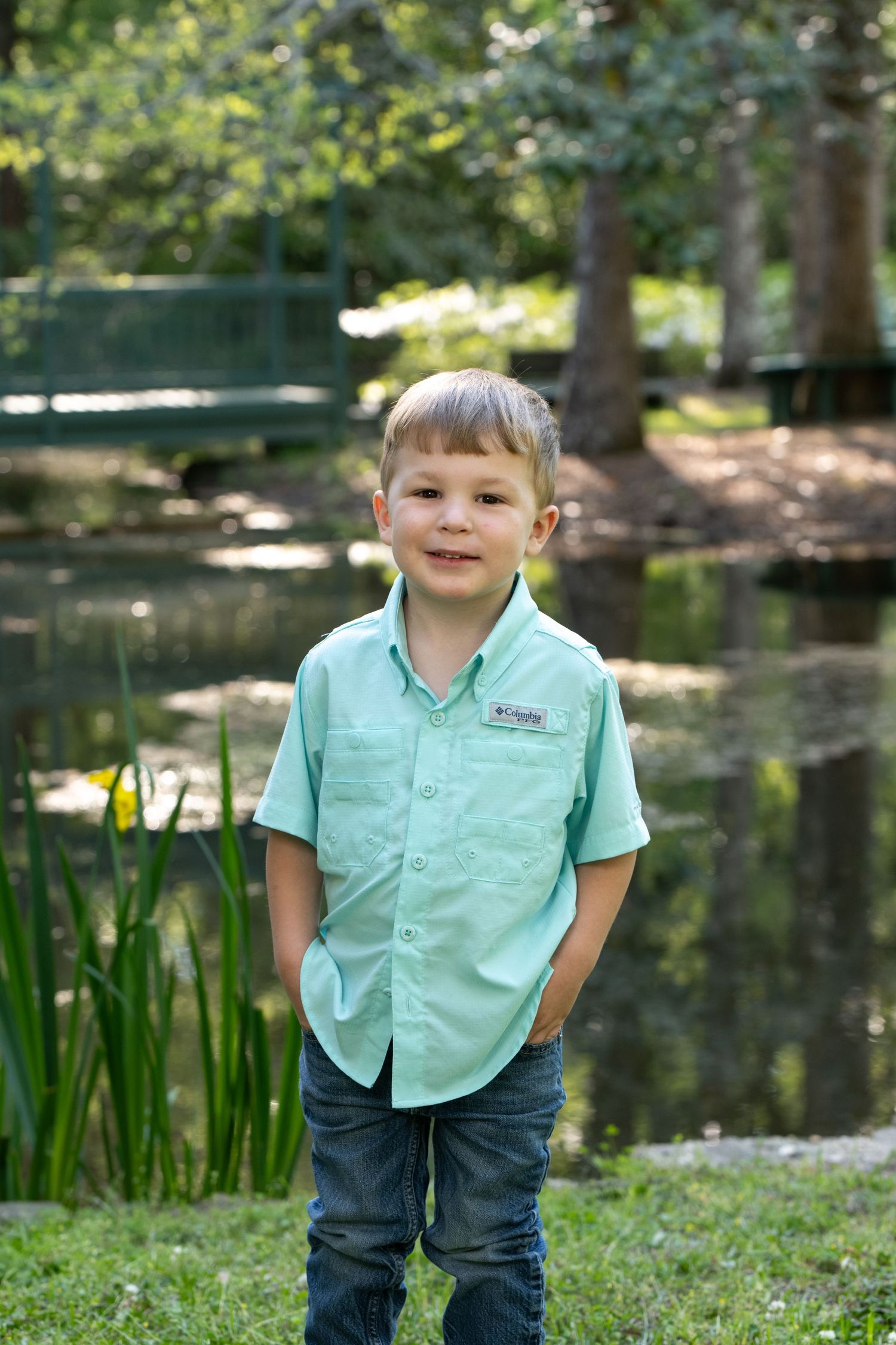 Young boy in mint green shirt and jeans smiling outdoors near a pond with lush green trees.