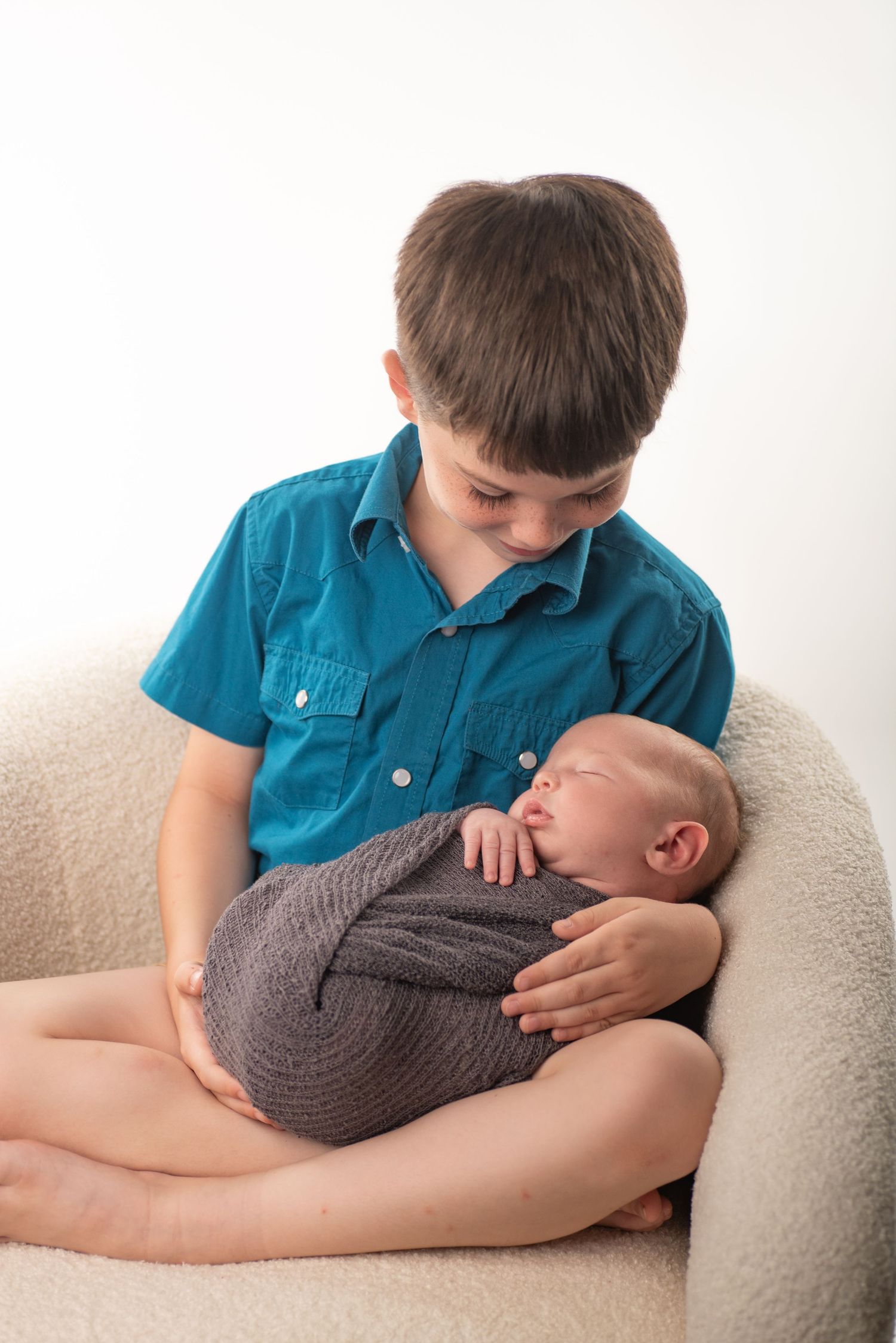 Young boy in teal shirt gently holding sleeping newborn baby wrapped in gray swaddle on white chair.