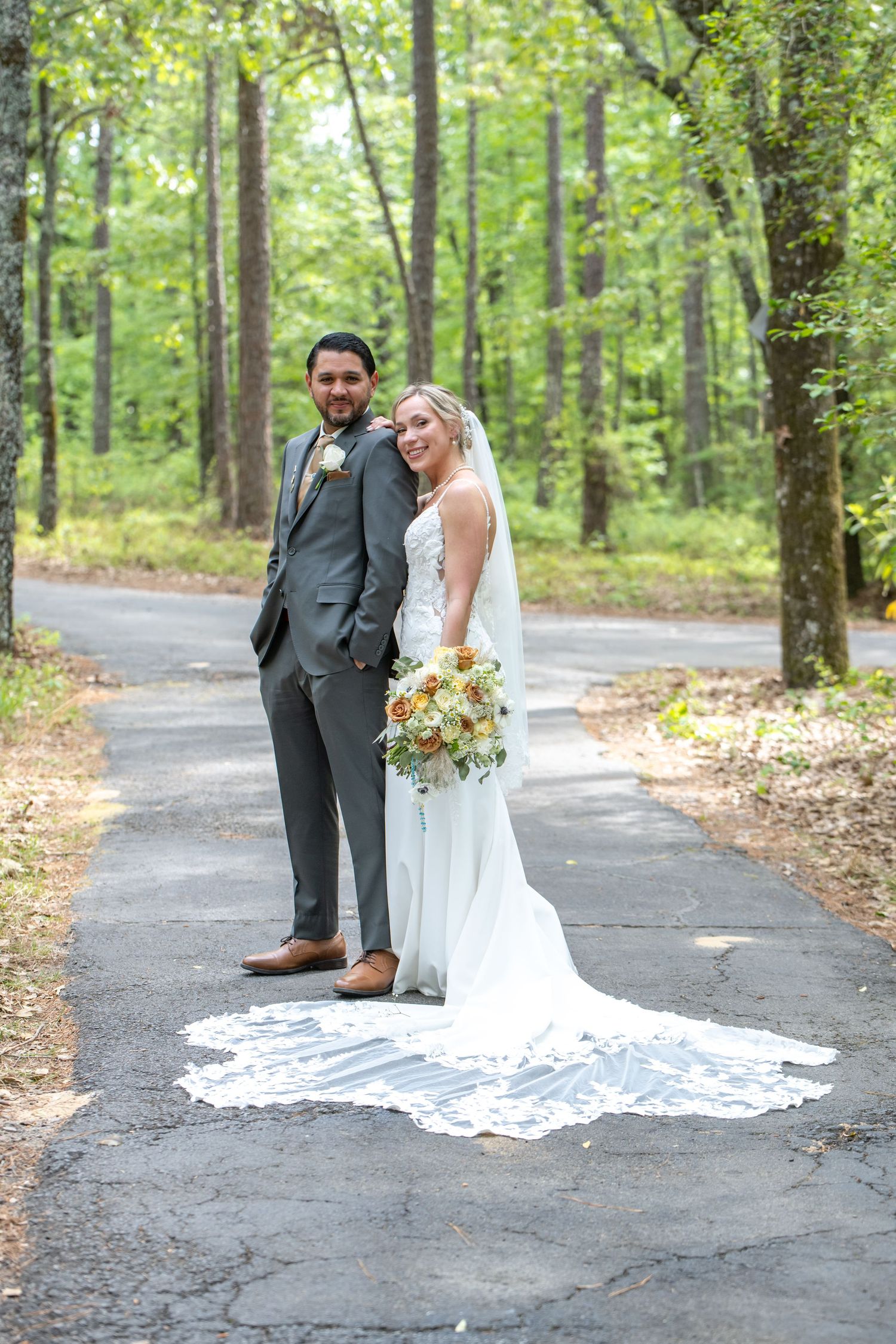 Bride and groom pose on a tree-lined path, she in a white gown with long train, he in a gray suit.