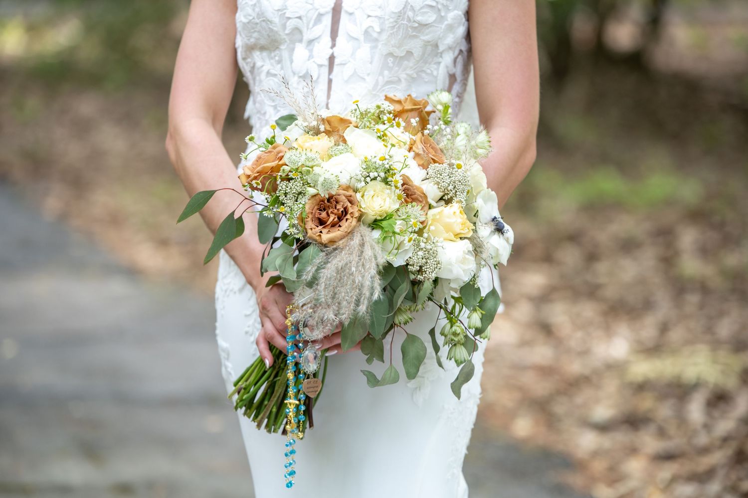 Bride holding a lush wedding bouquet with white, peach, and gold flowers mixed with eucalyptus greenery outdoors.