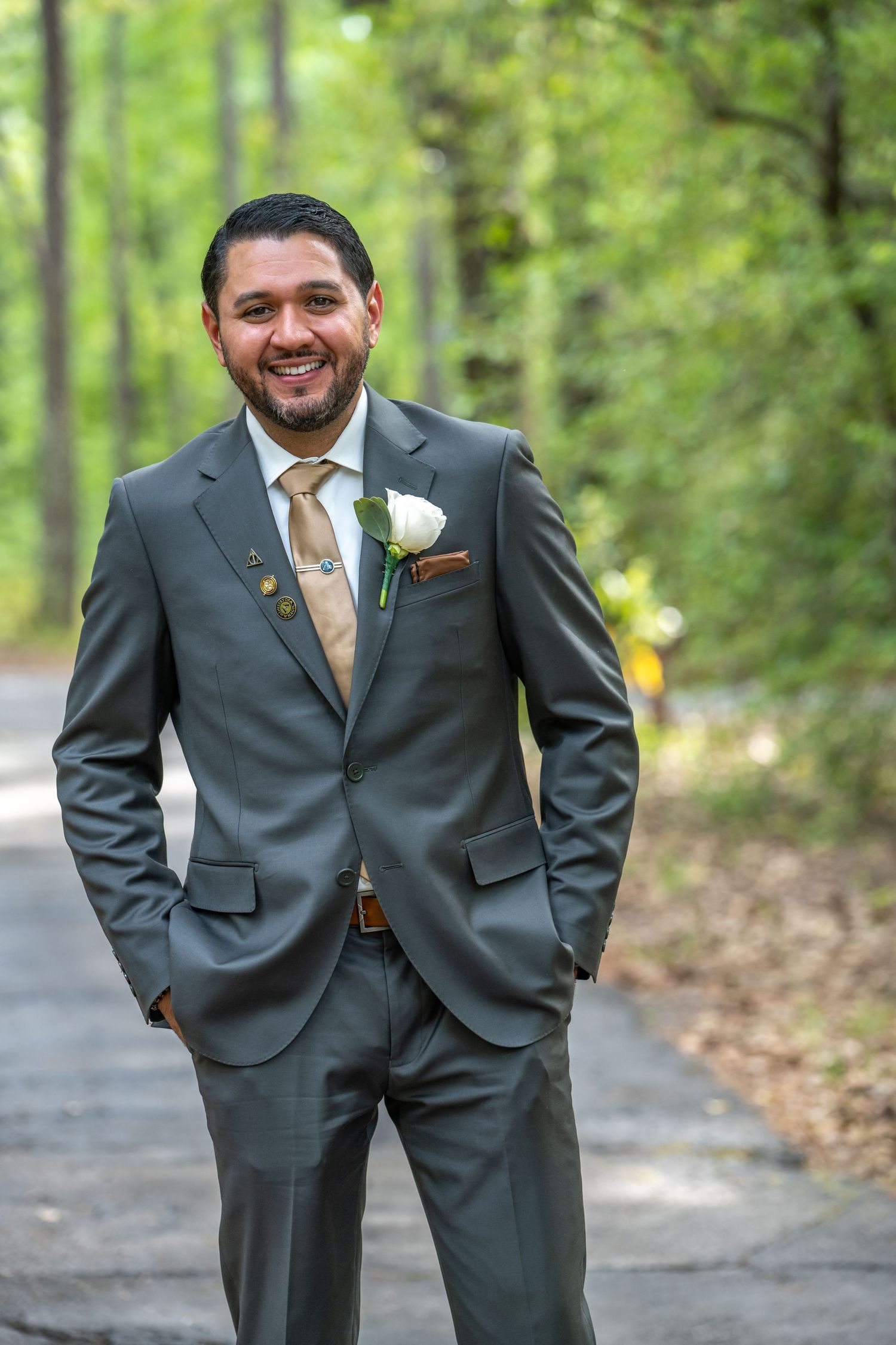 Smiling groom in gray suit with white boutonniere and tan tie poses outdoors among green trees.