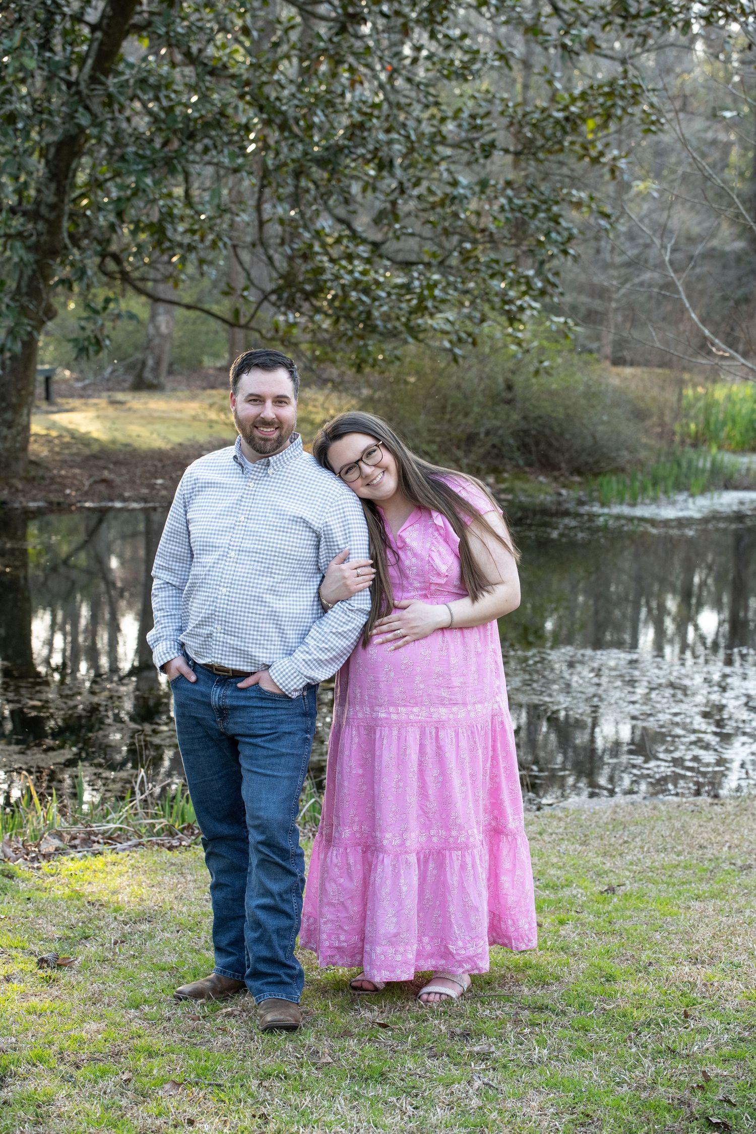 A couple poses together by a pond on a grassy area with trees in the background.
