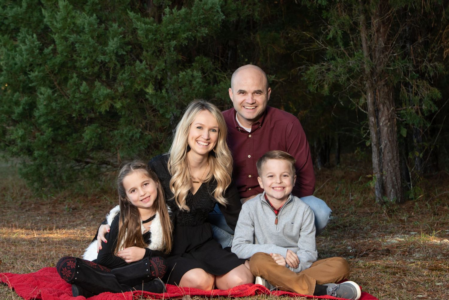 Family of four sitting on a red blanket for outdoor portrait session in wooded setting.
