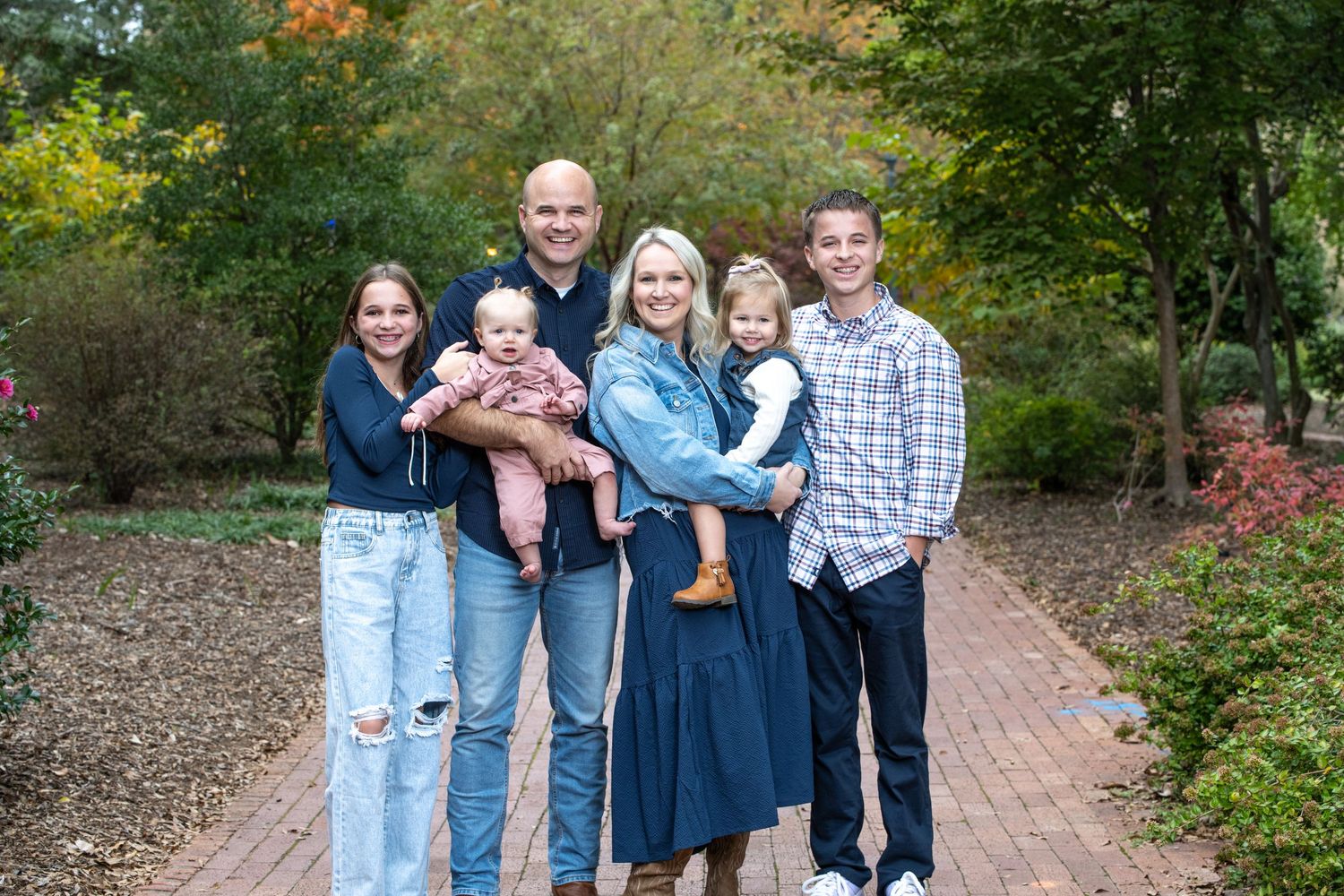 Happy family of six posing outdoors on a brick path surrounded by lush green autumn trees.