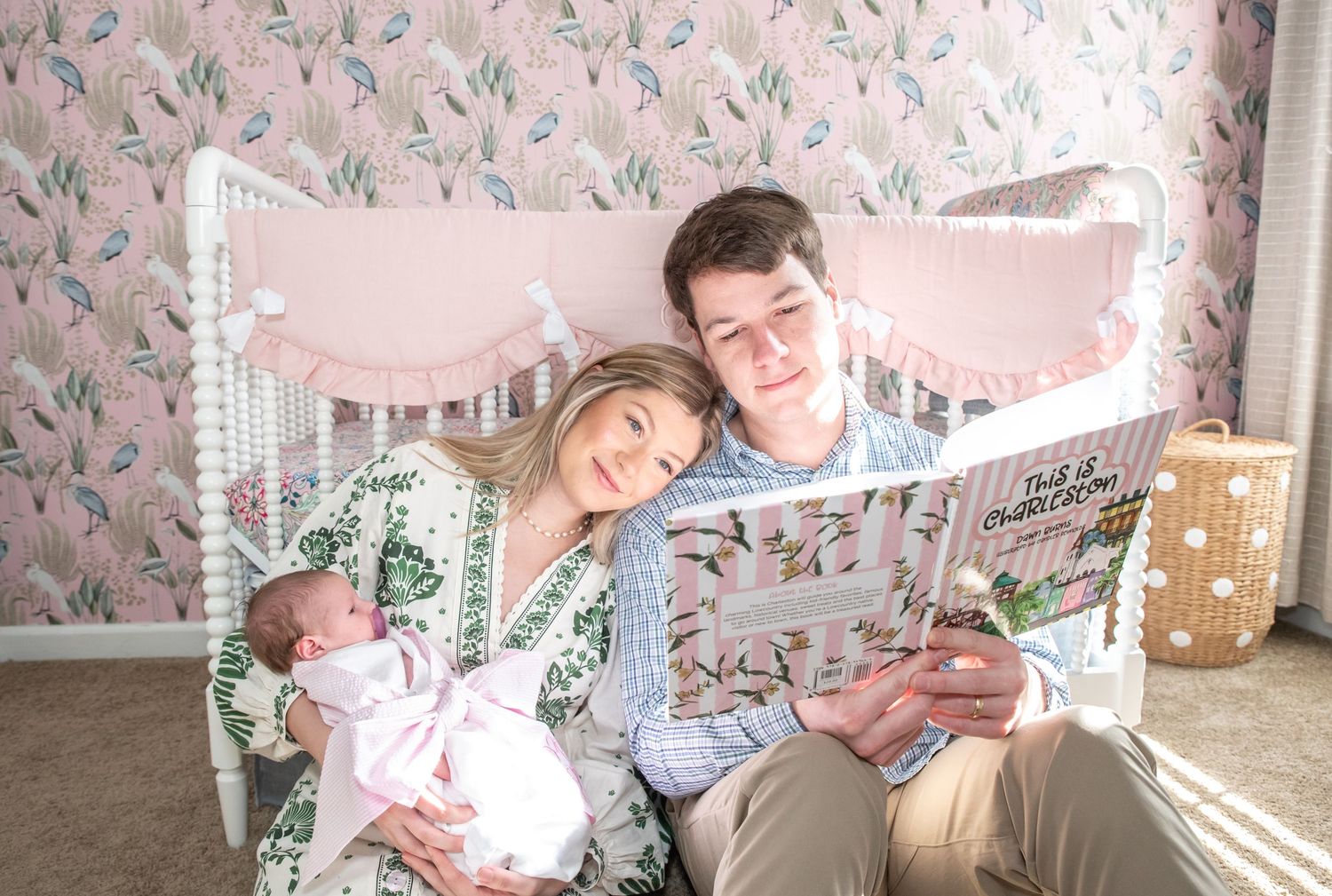 Couple sitting by white crib reading to newborn baby in floral nursery room.