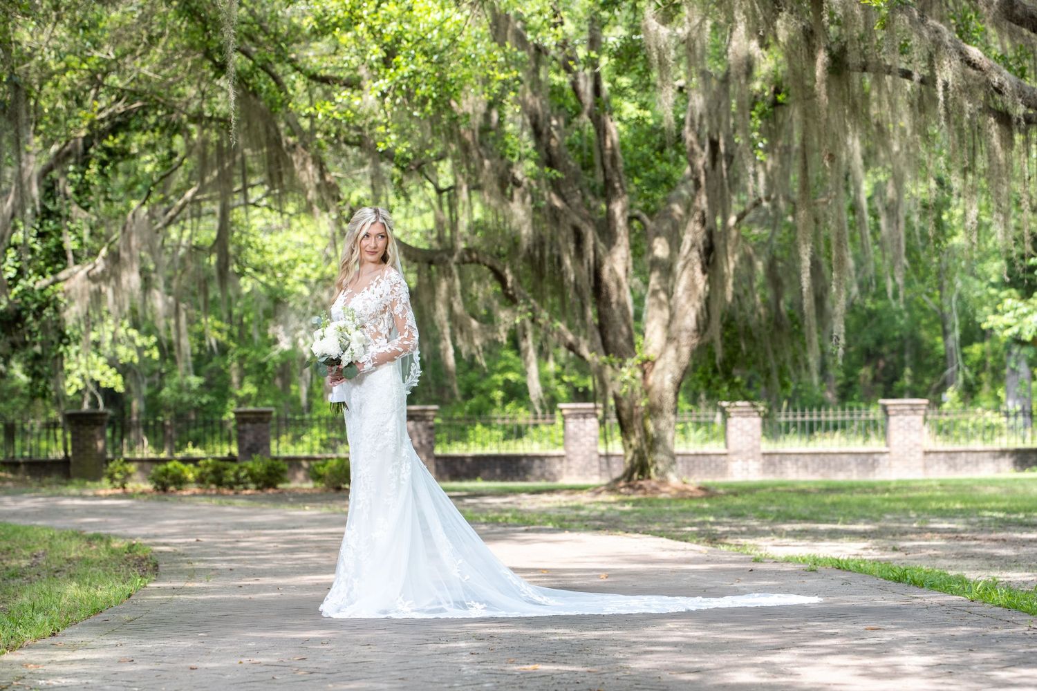 A scenic wedding photo taken in a lush garden with Spanish moss draped trees and stone columns.