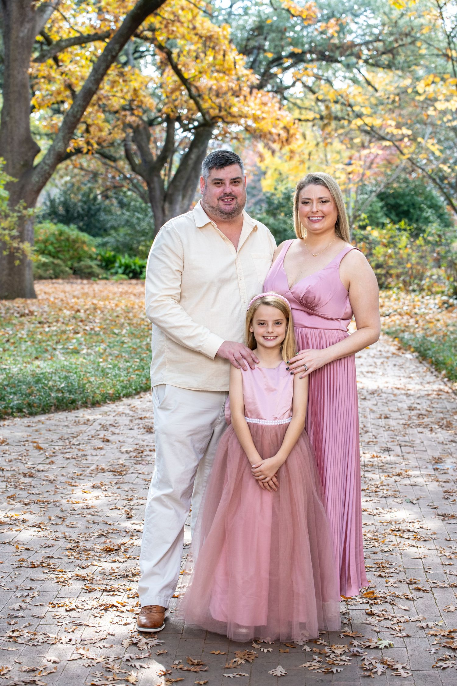 Family of three posing in autumn park, parents in pink and white outfits with young daughter in pink tulle dress.