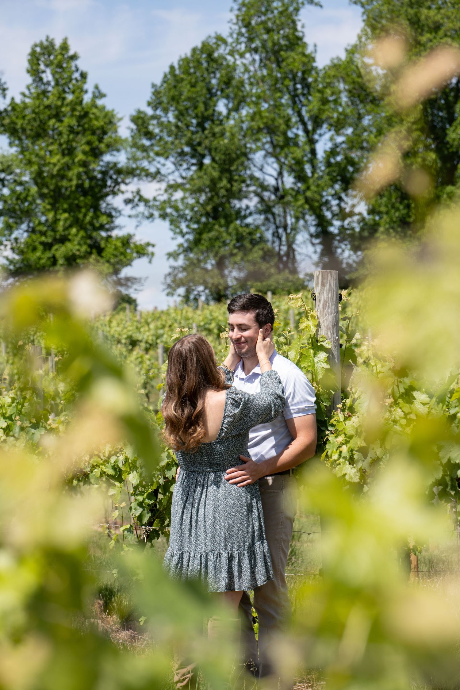 A couple embraces and smiles at each other among lush green vineyard rows on a sunny day.