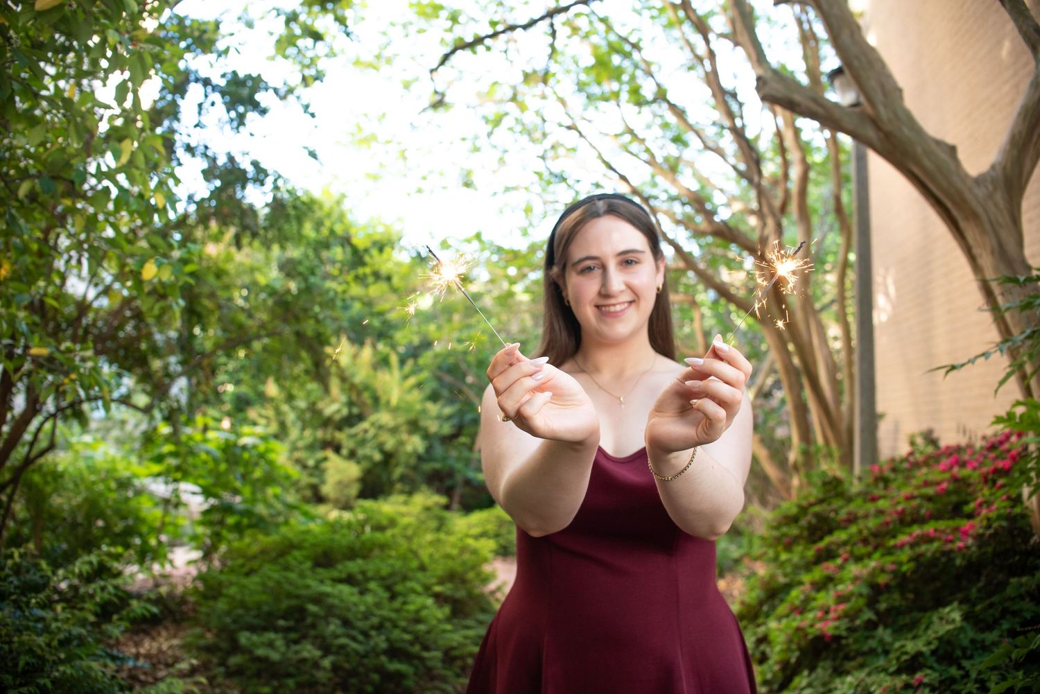 Young woman in burgundy dress holding sparklers outdoors, smiling in a lush green garden setting.