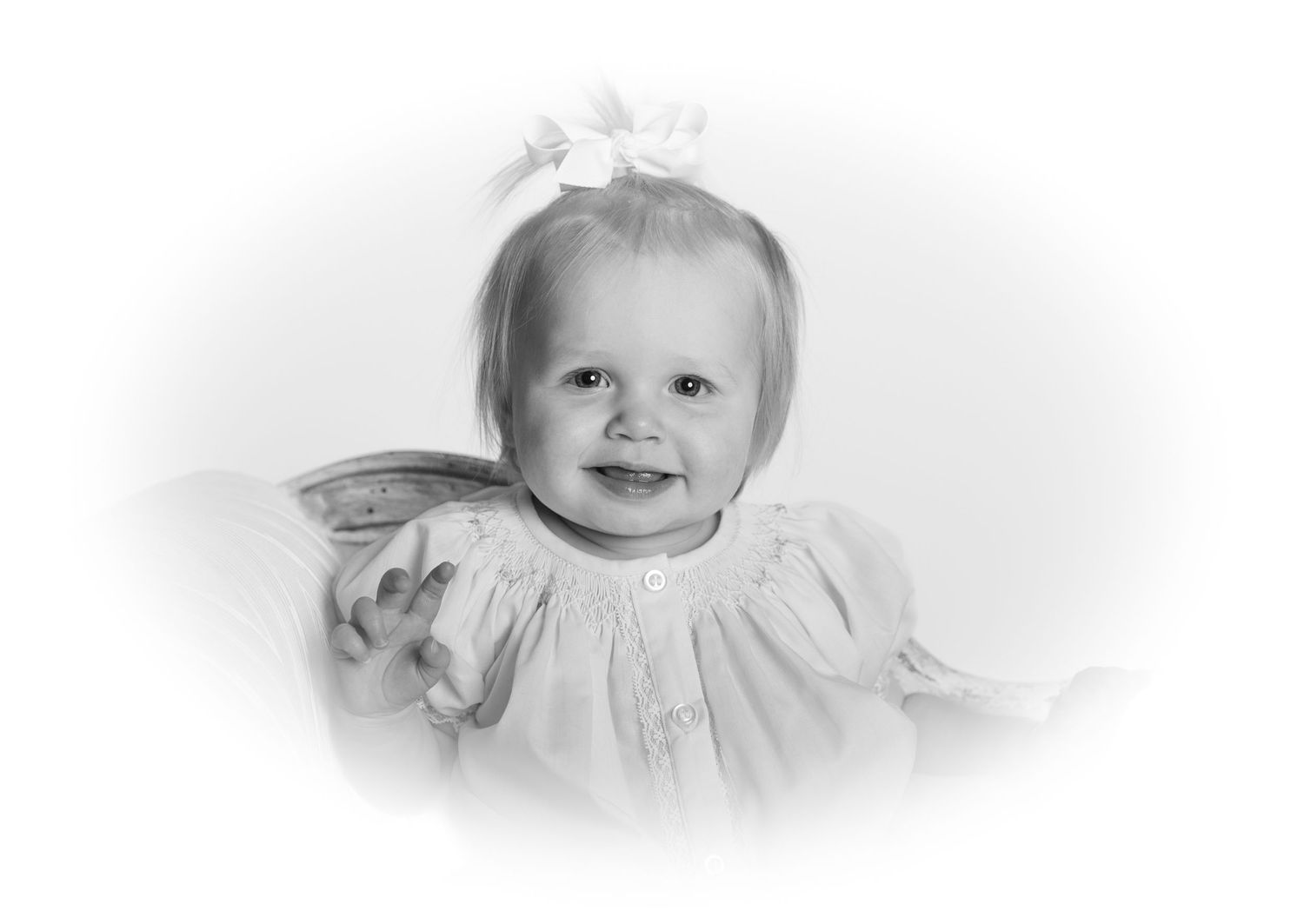 Black and white portrait of a smiling baby girl wearing a white dress and bow, classic studio photography.