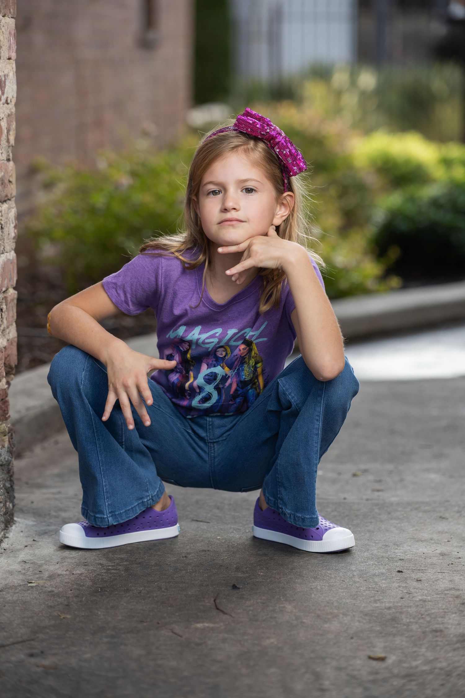 Young girl in purple shirt and jeans crouching outdoors, wearing pink headband and purple sneakers.