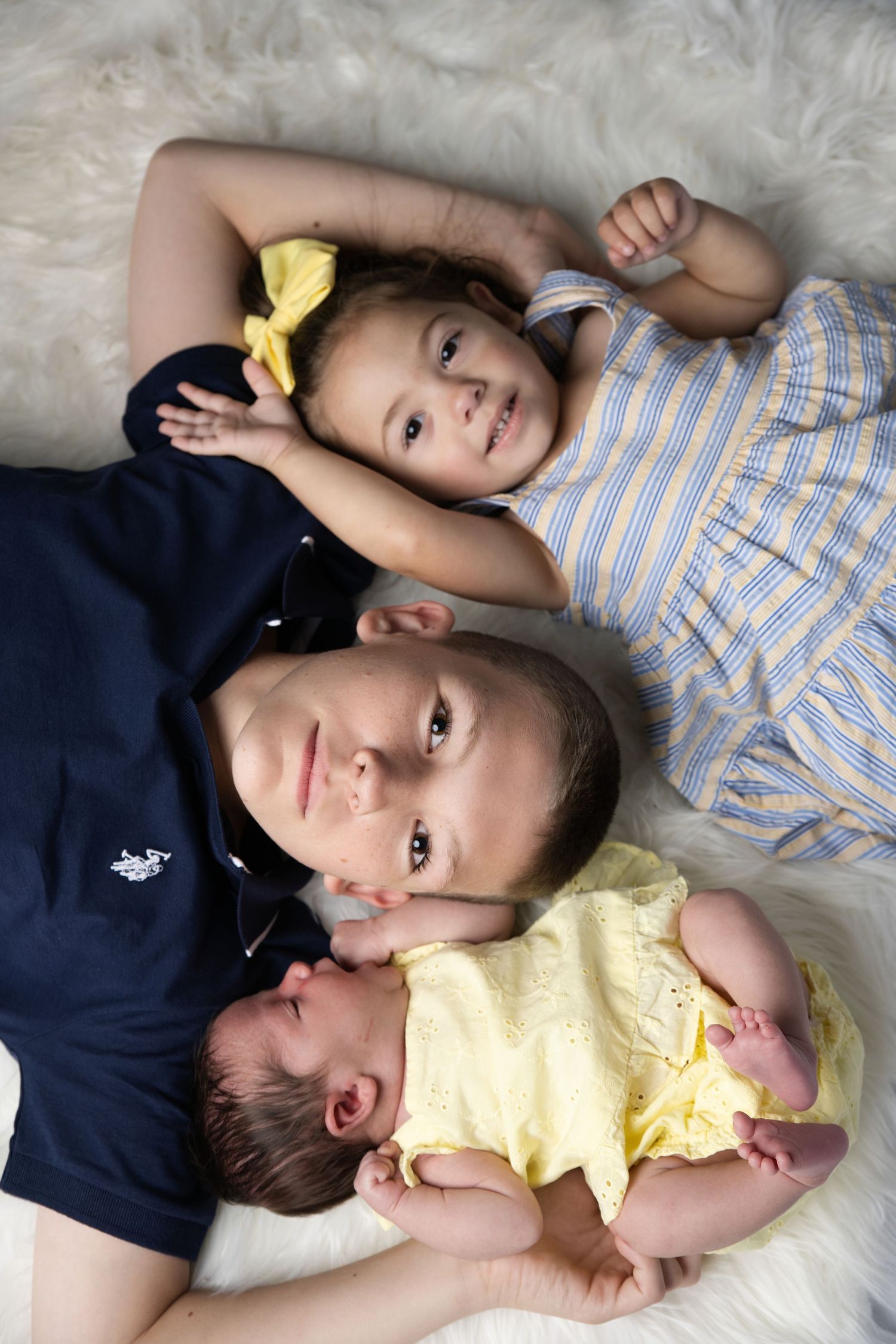 Three siblings lying together on a fluffy white rug, including a newborn in yellow, a boy, and a girl with a bow.