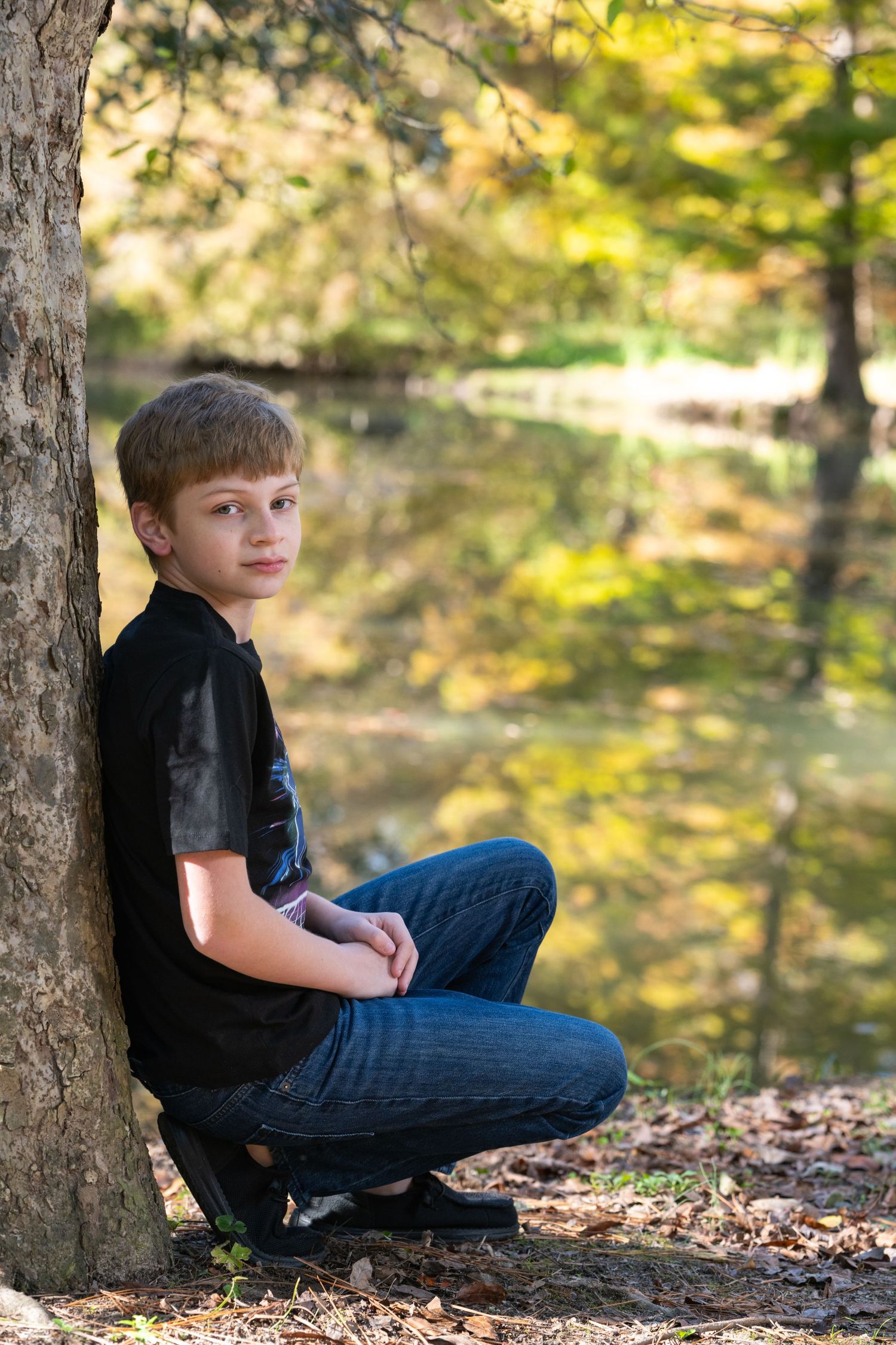 A teenage boy in a black shirt and jeans sits against a tree by a reflective pond in an autumn forest.