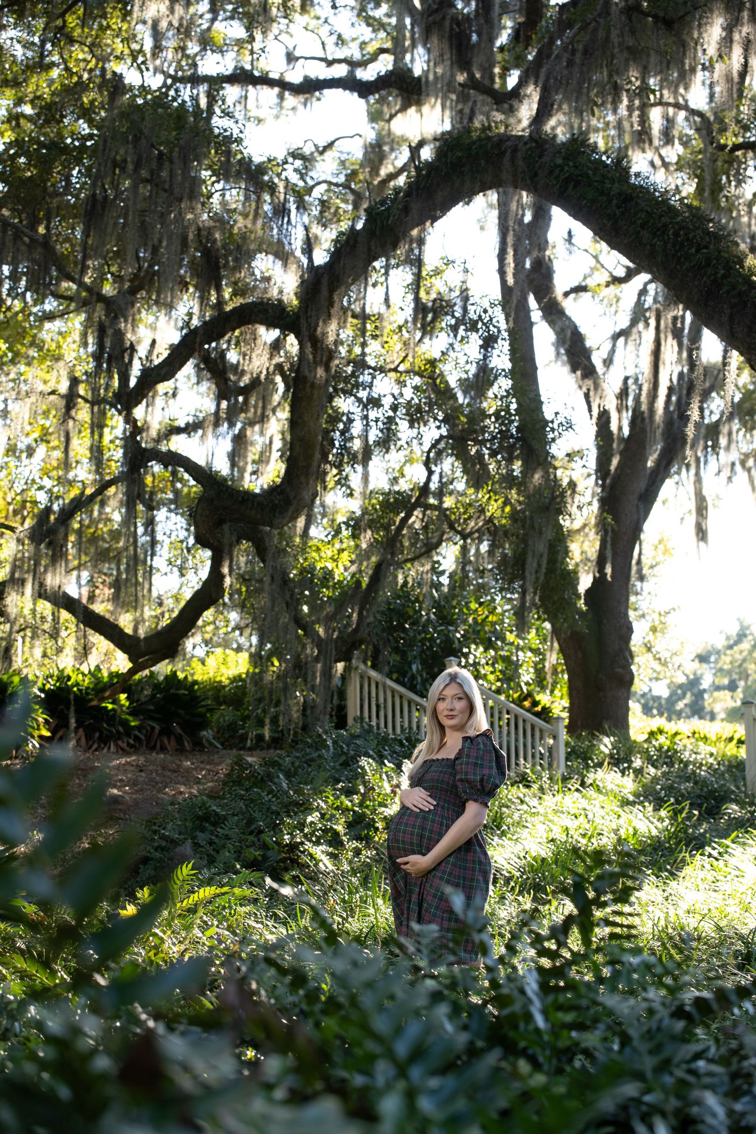 Pregnant woman in plaid dress standing under moss-draped oak trees in a sunlit garden setting.