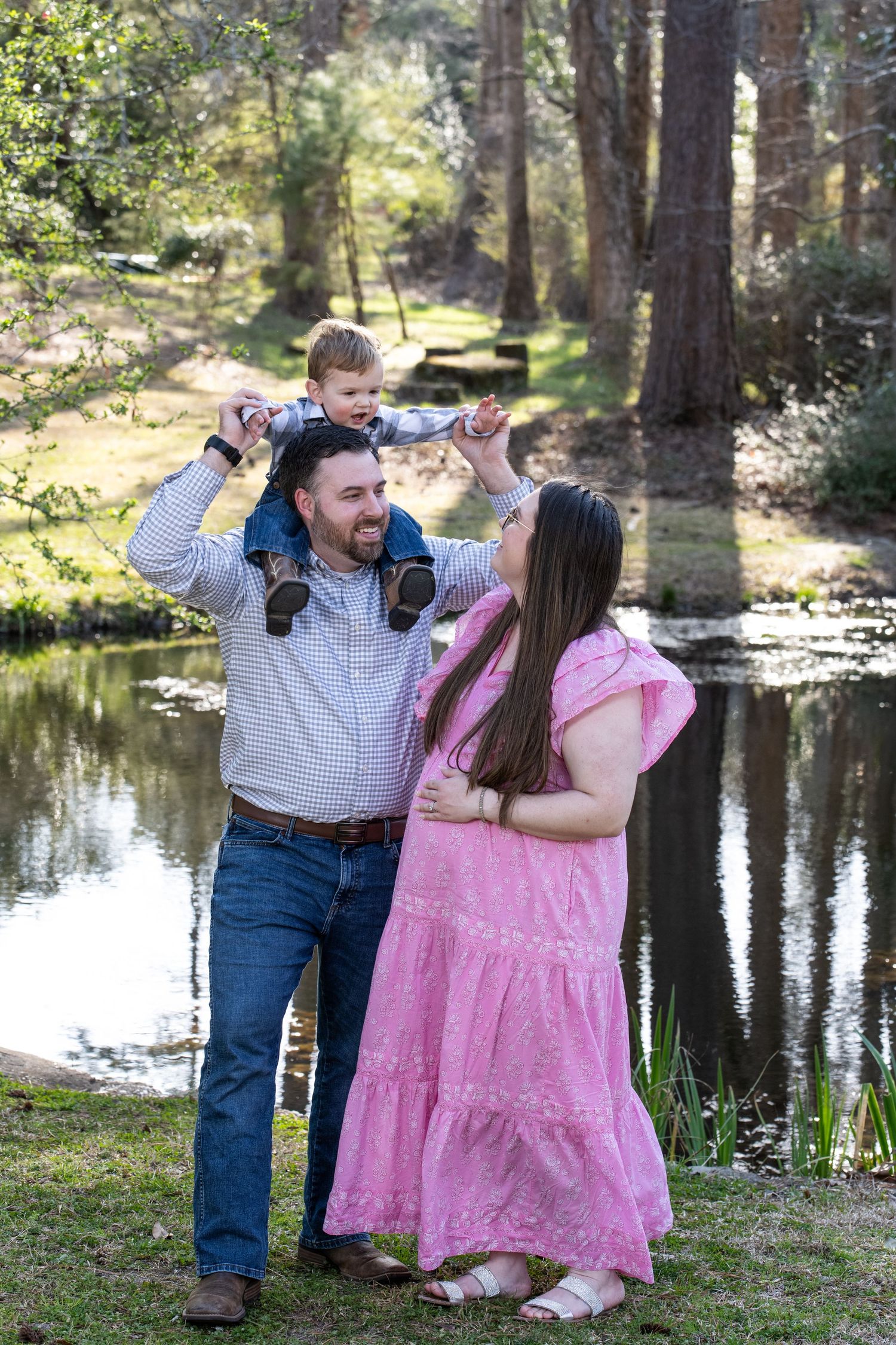 Family enjoying a tender moment together by a reflective pond in a sunlit wooded area during springtime.