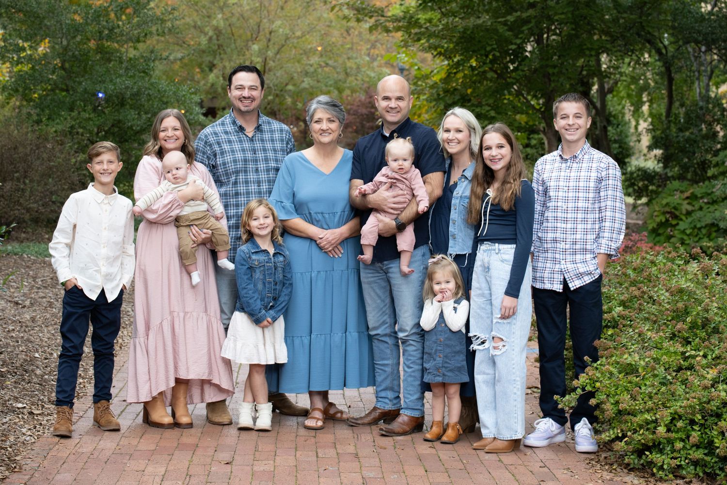 Large family group portrait smiling outdoors on a brick path surrounded by lush green trees in autumn.