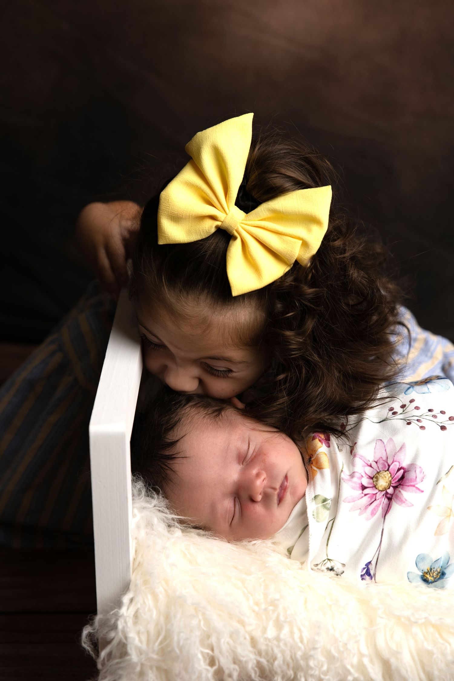 Young girl with yellow bow kisses sleeping newborn baby wrapped in floral swaddle in white box.
