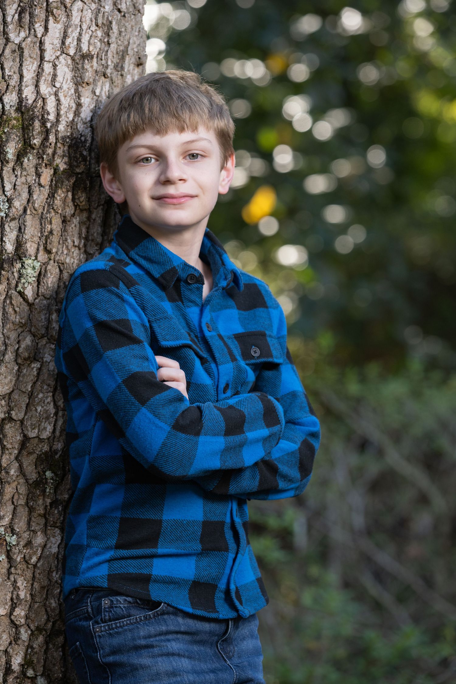 A young boy in a blue plaid flannel shirt leans against a tree with arms crossed, smiling outdoors.
