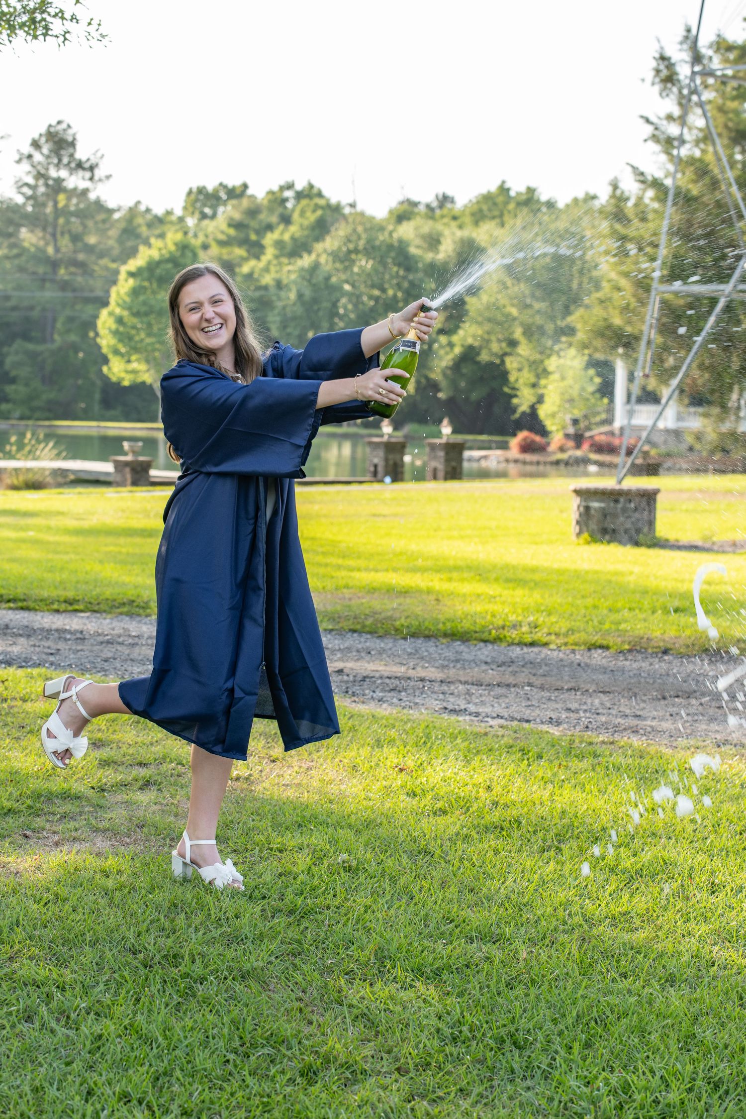 Woman in blue graduation gown joyfully spraying champagne outdoors on green lawn with trees in background.