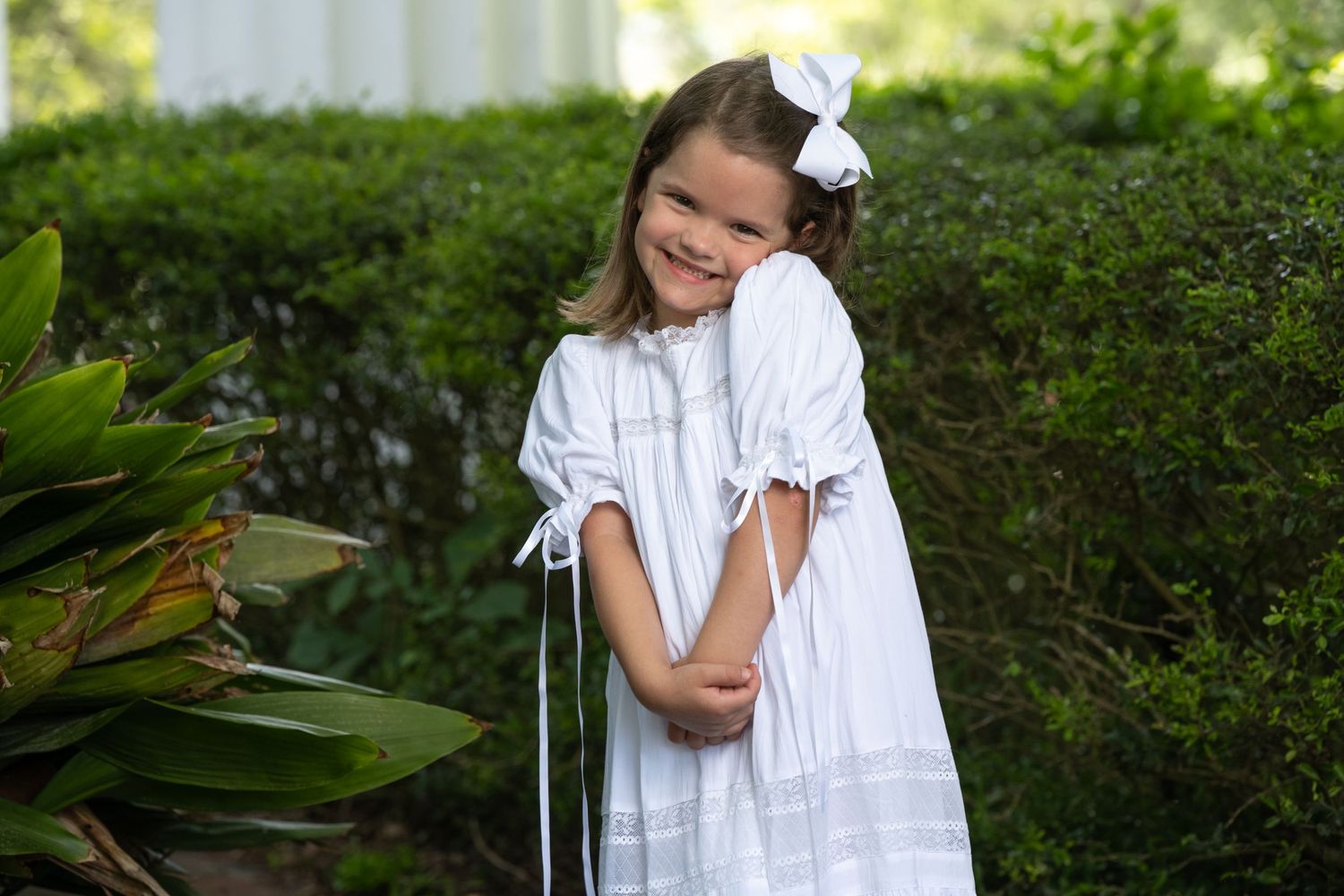 Young girl in white dress and bow smiles shyly in garden setting.