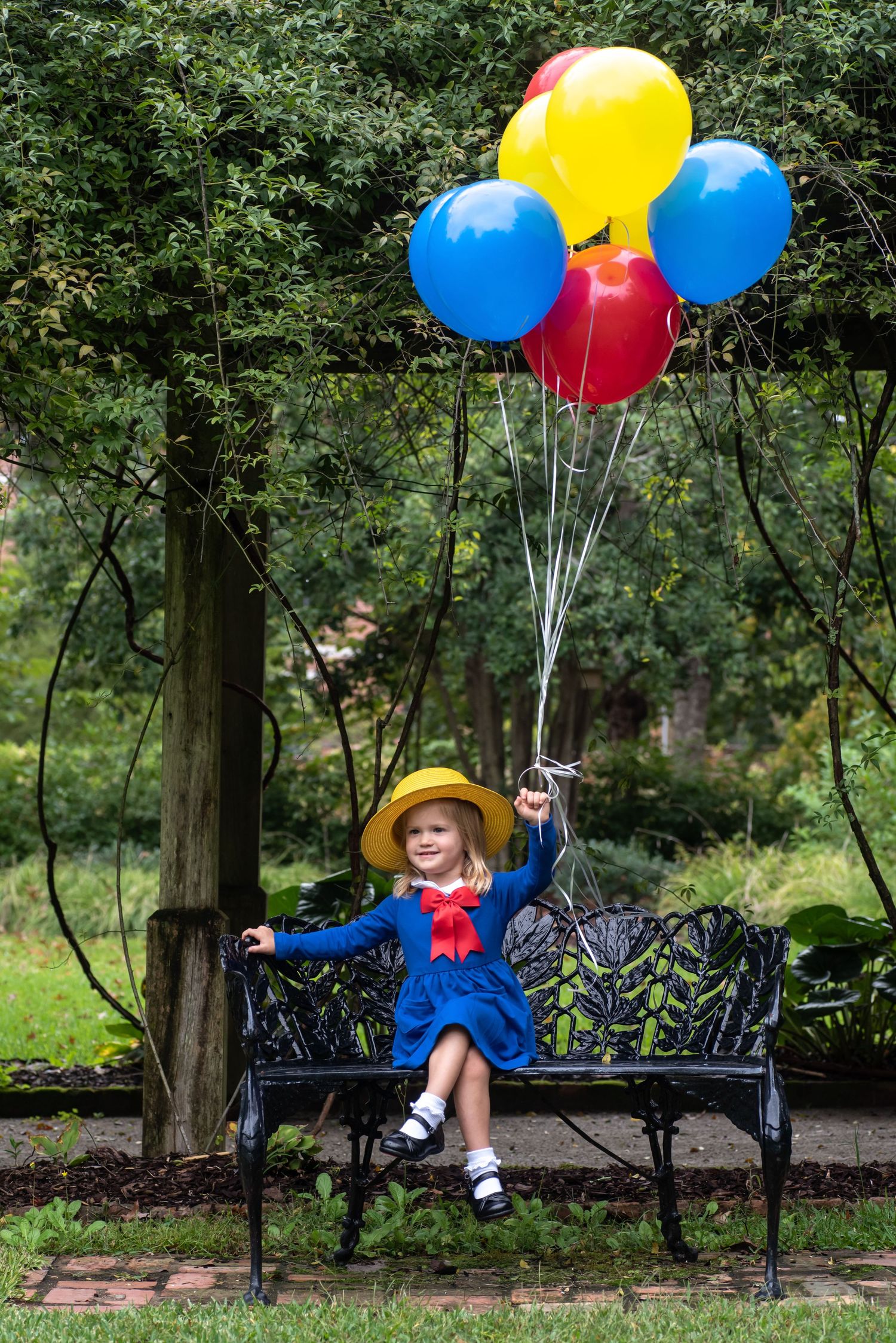 Child dressed as Madeline holding colorful balloons while sitting on wrought iron bench in a garden.
