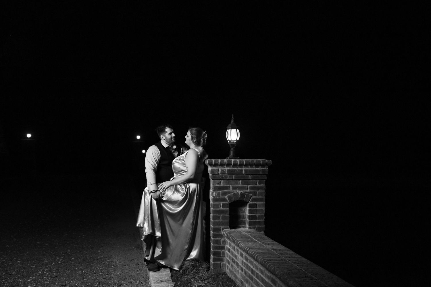 A couple stands in intimate embrace near a brick wall with lamp post at night, captured in black and white.