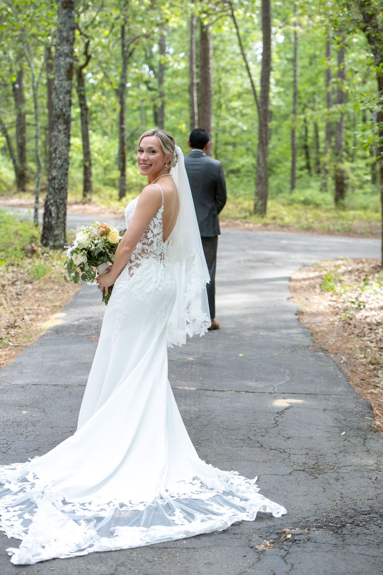 Bride in white lace gown with veil holds bouquet on forest path, smiling back as groom walks ahead.