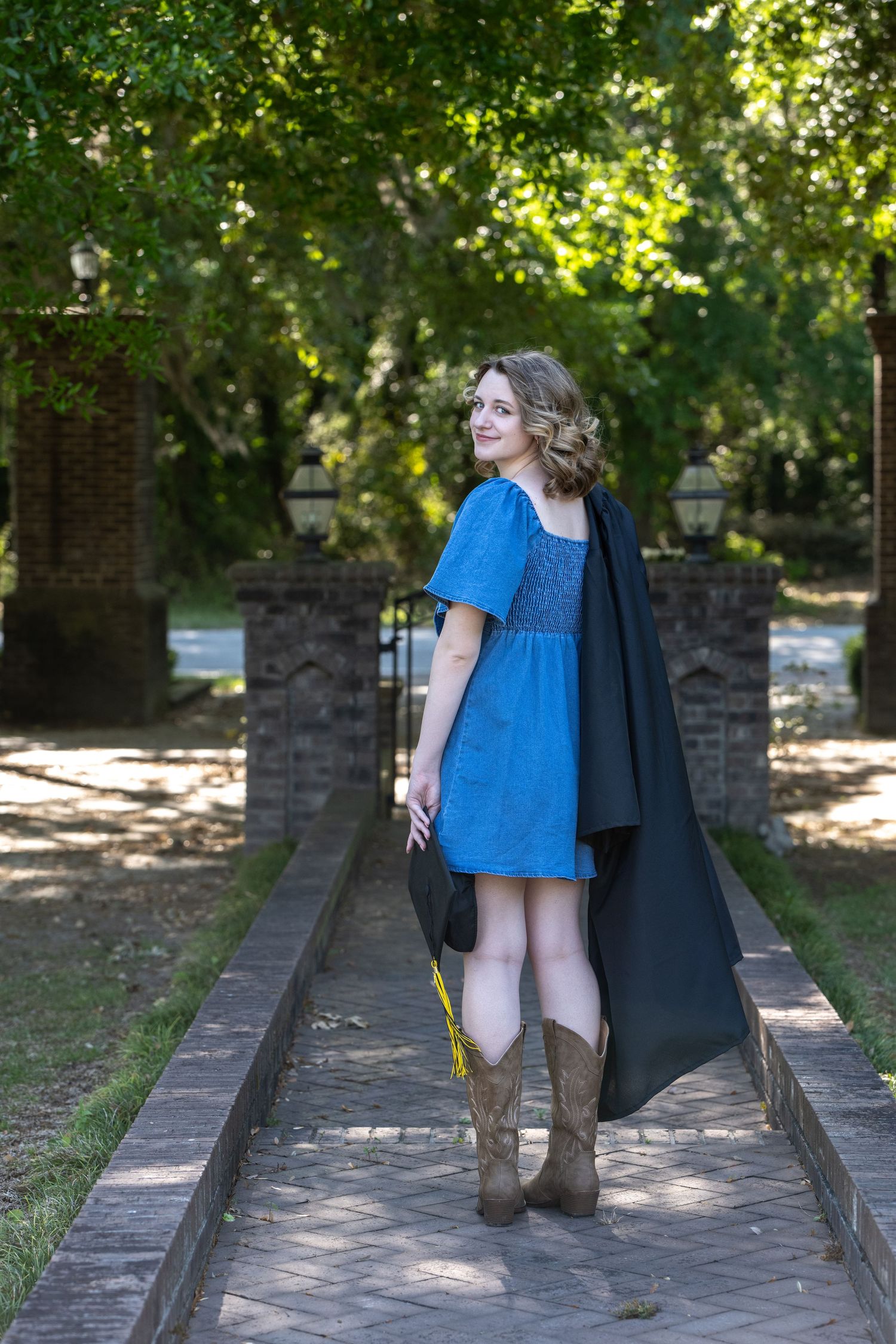 Young woman in blue dress and cowboy boots wearing graduation gown holds cap outdoors on brick path.