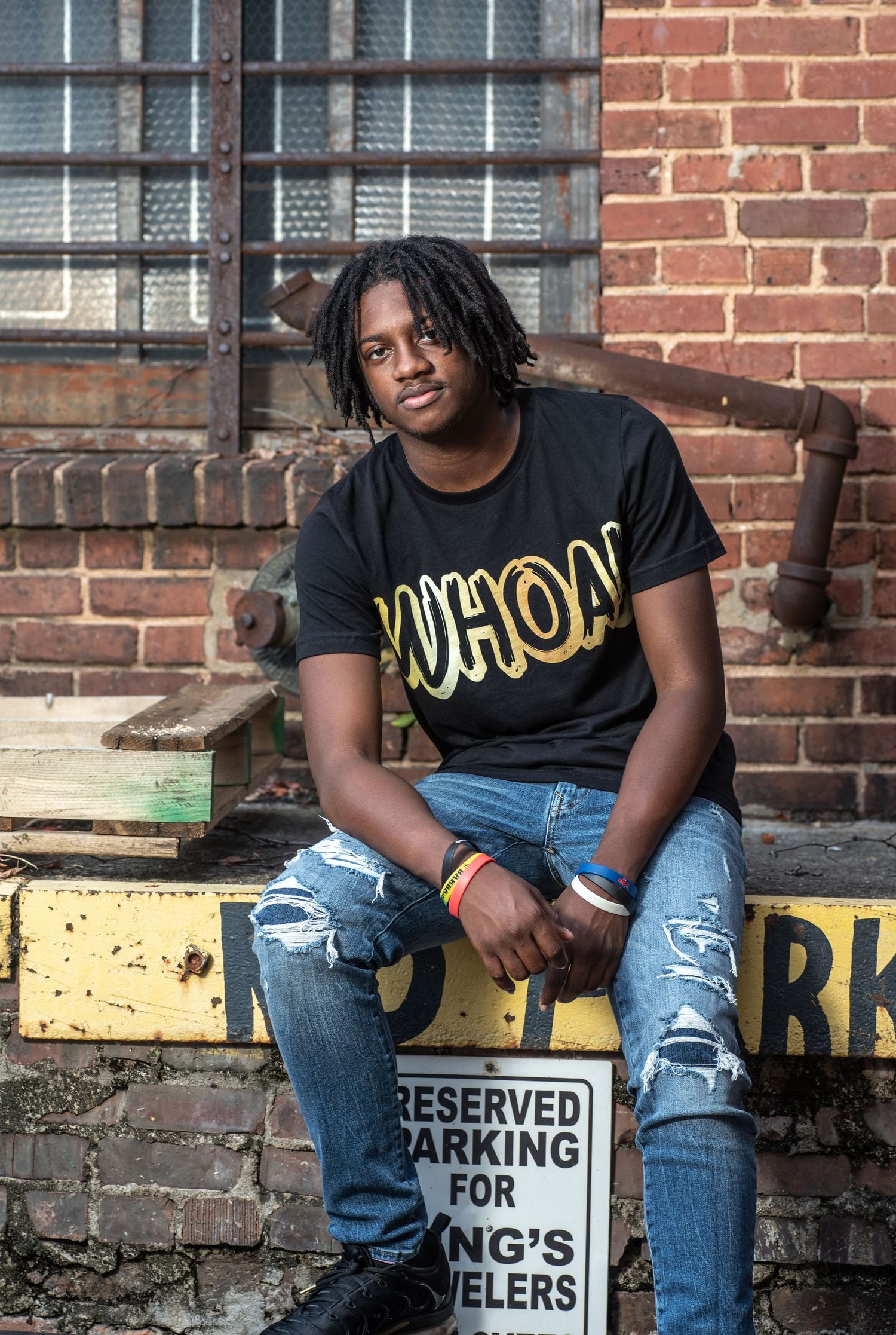 Person in a 'WHOA!' t-shirt and ripped jeans sits casually on a yellow barrier near a brick wall.