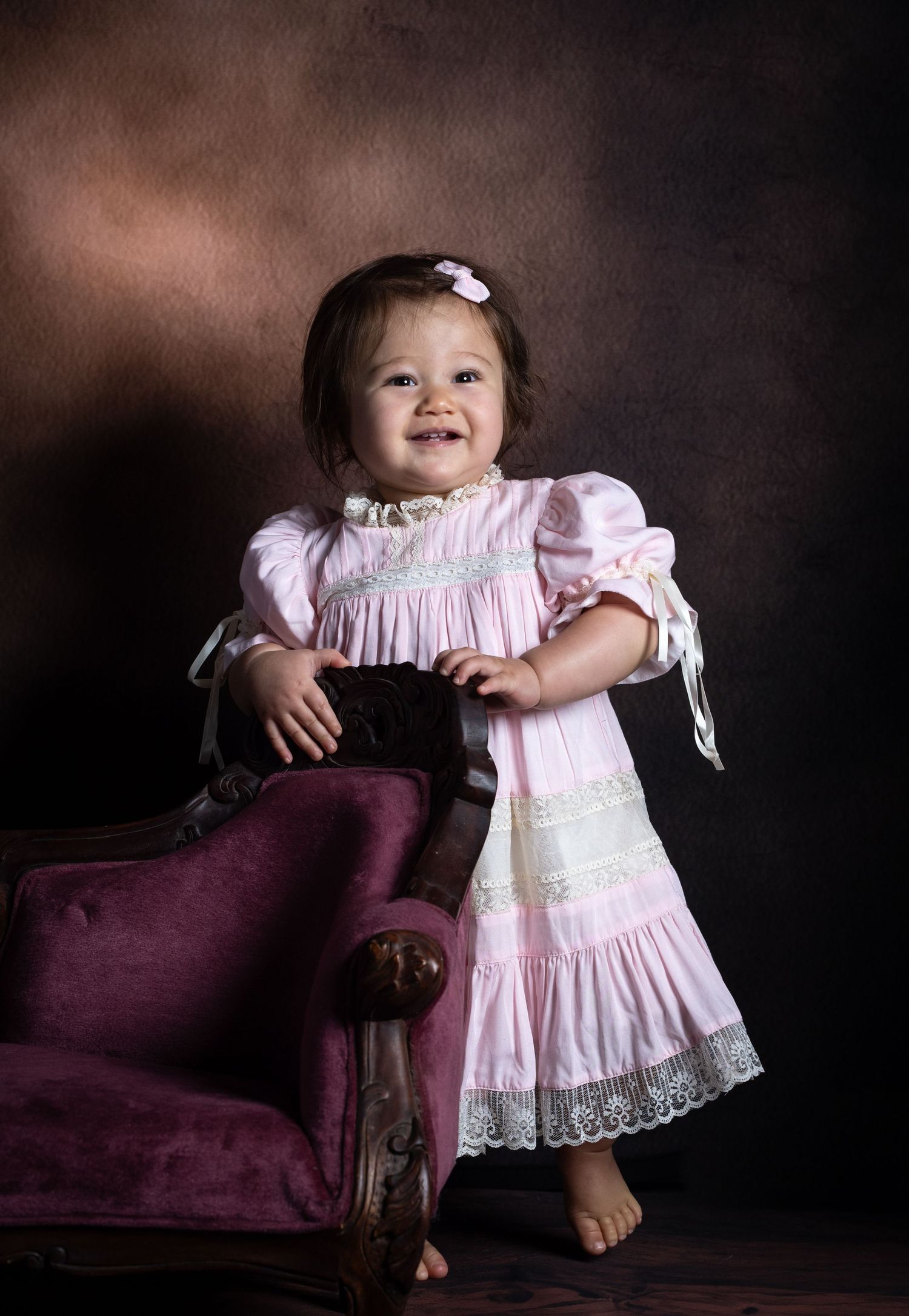 Smiling toddler girl in pink lace dress standing beside vintage purple chair in professional studio portrait.