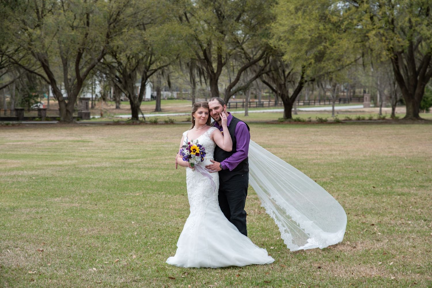 Two people share a romantic moment in their wedding attire on a grassy field surrounded by trees.