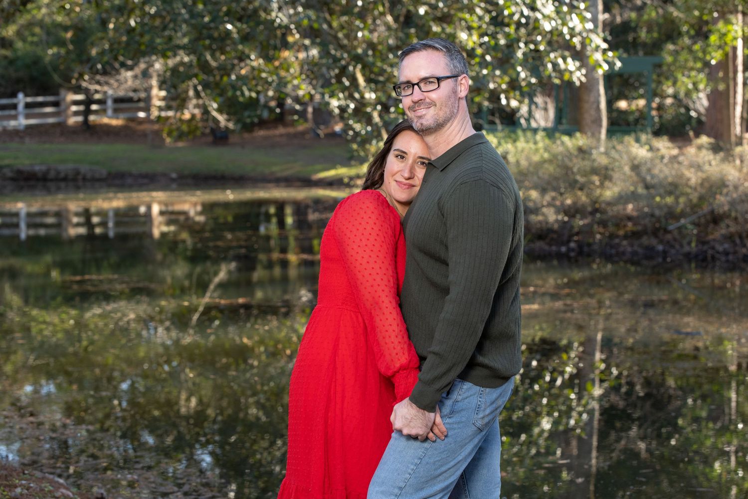 Couple embracing outdoors near a pond, woman in red dress and man in green sweater, surrounded by lush trees.