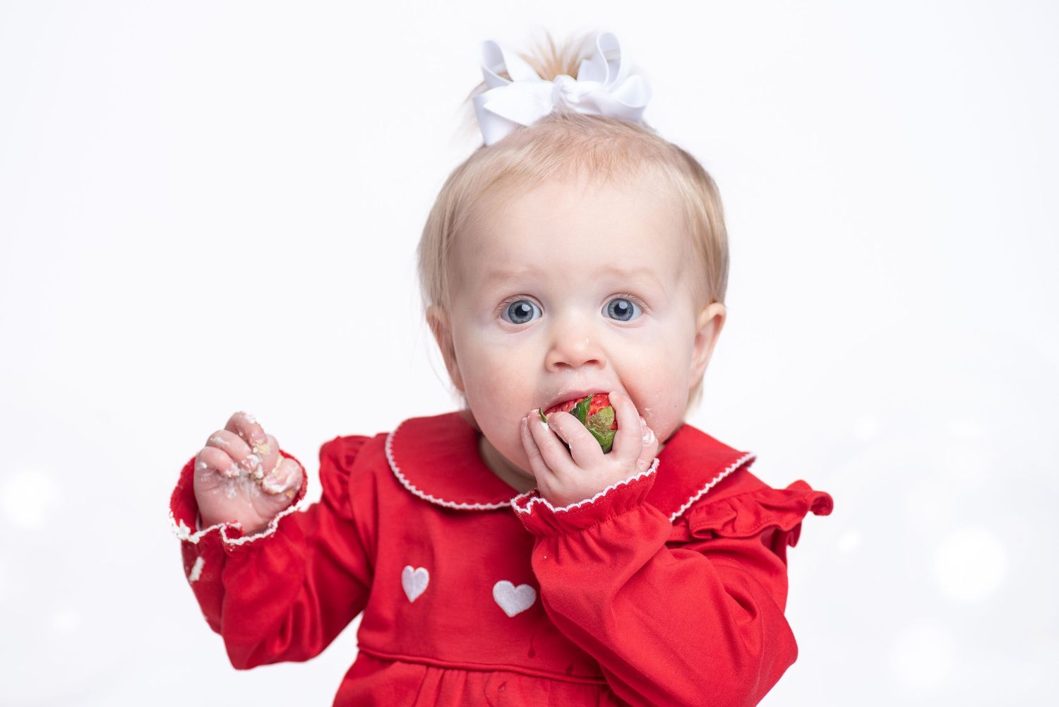 Cute baby girl in red dress with white bow eating a strawberry against a light background.