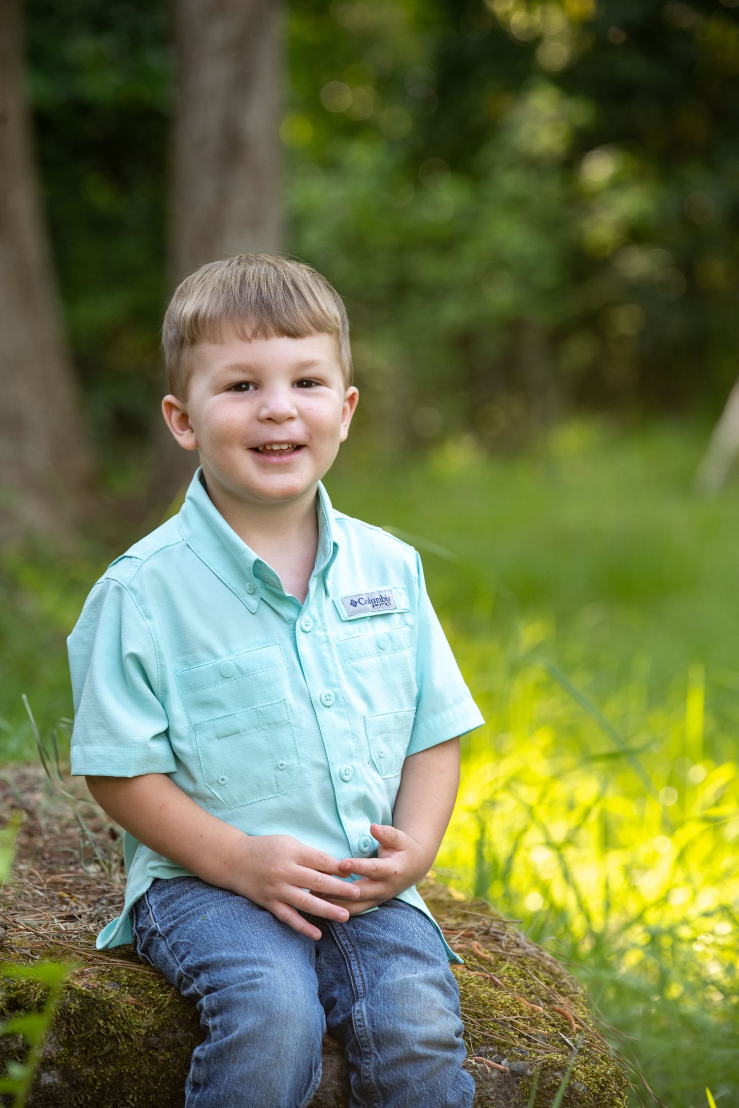 Smiling young boy in a light blue Columbia shirt and jeans sitting on a rock in a lush green outdoor setting.