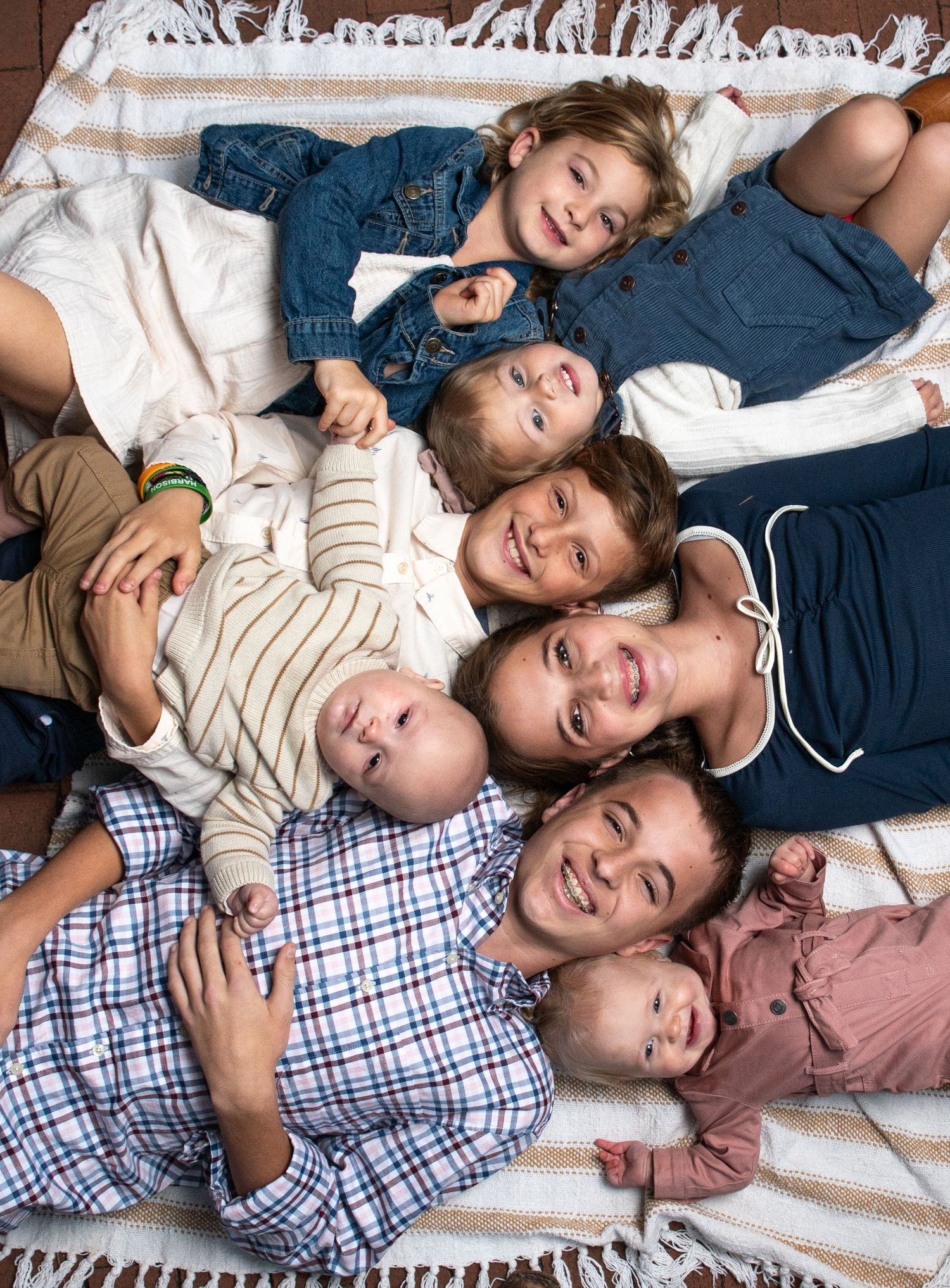 A joyful family portrait with two adults and five children lying together on a striped blanket, smiling up at the camera.