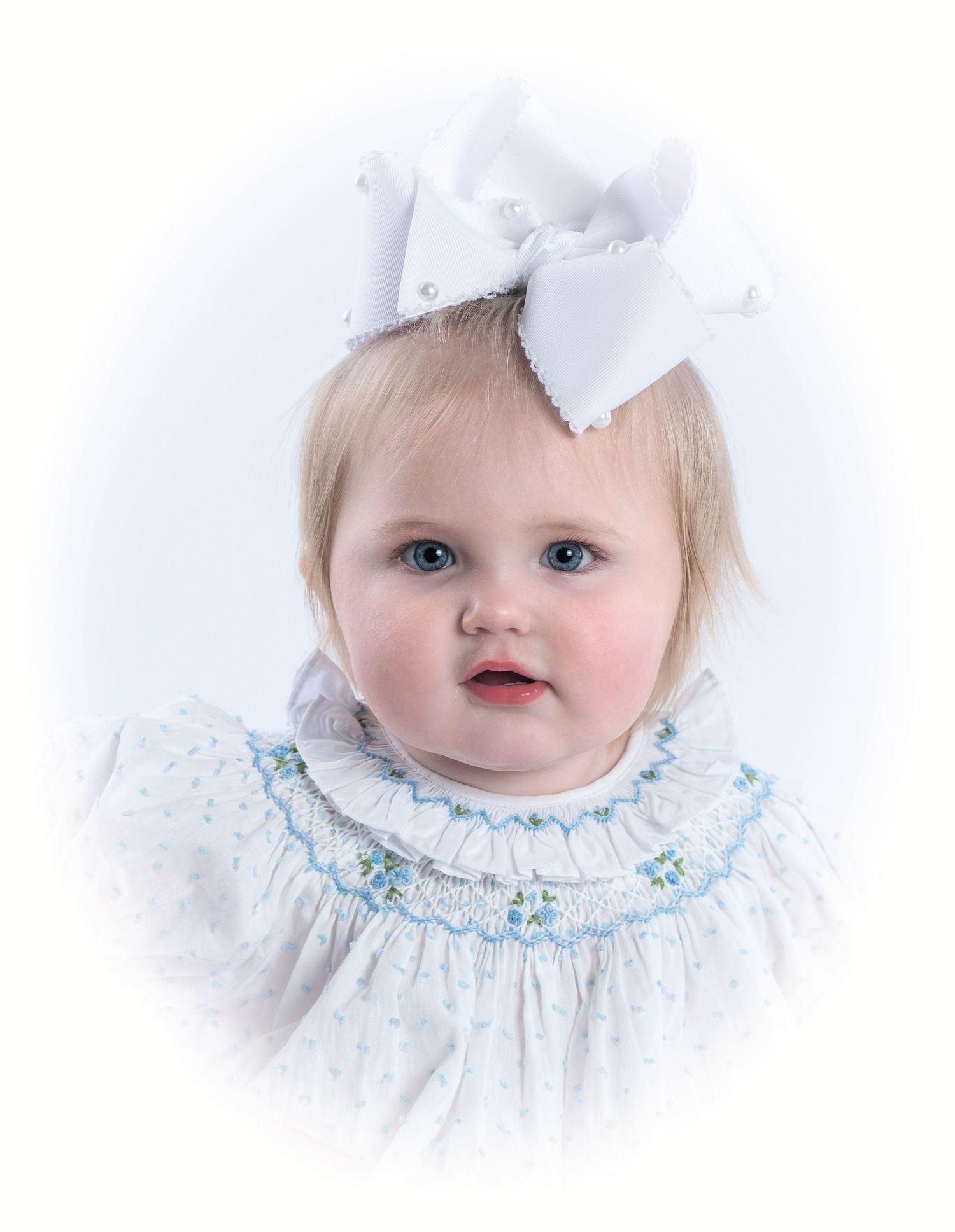 A small child wearing a white smocked dress with blue embroidery and a large white bow hair accessory in a soft portrait.