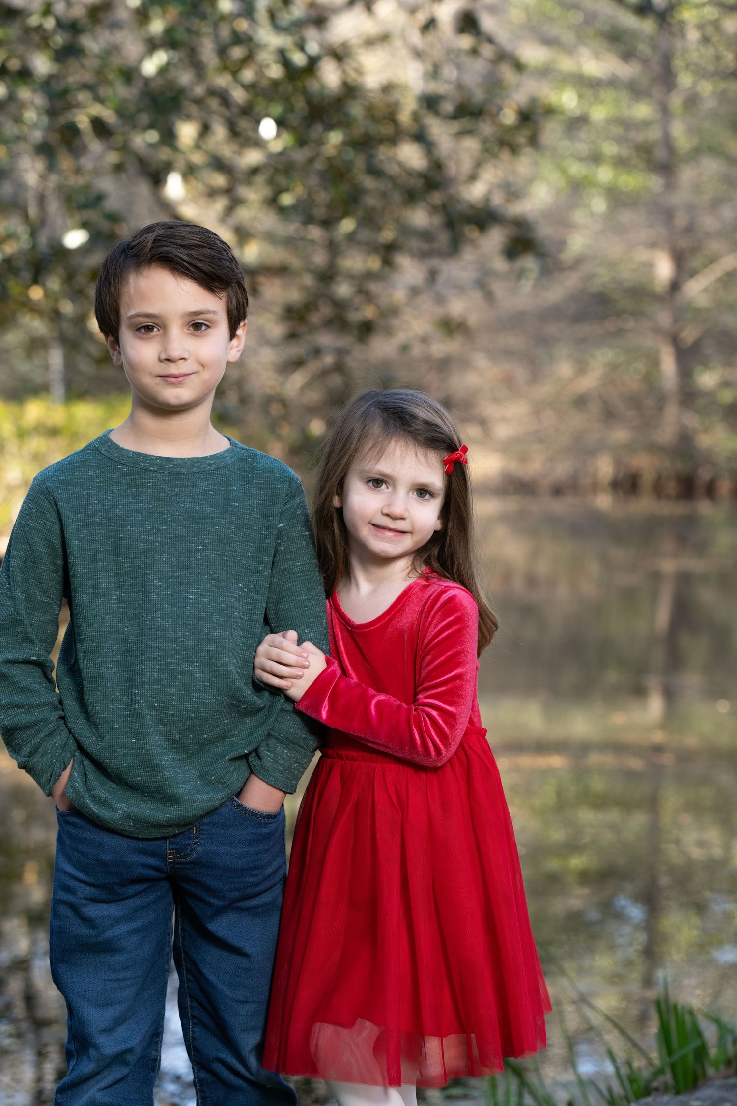 A boy in a green shirt and girl in a red dress pose together outdoors near a pond for a holiday portrait.