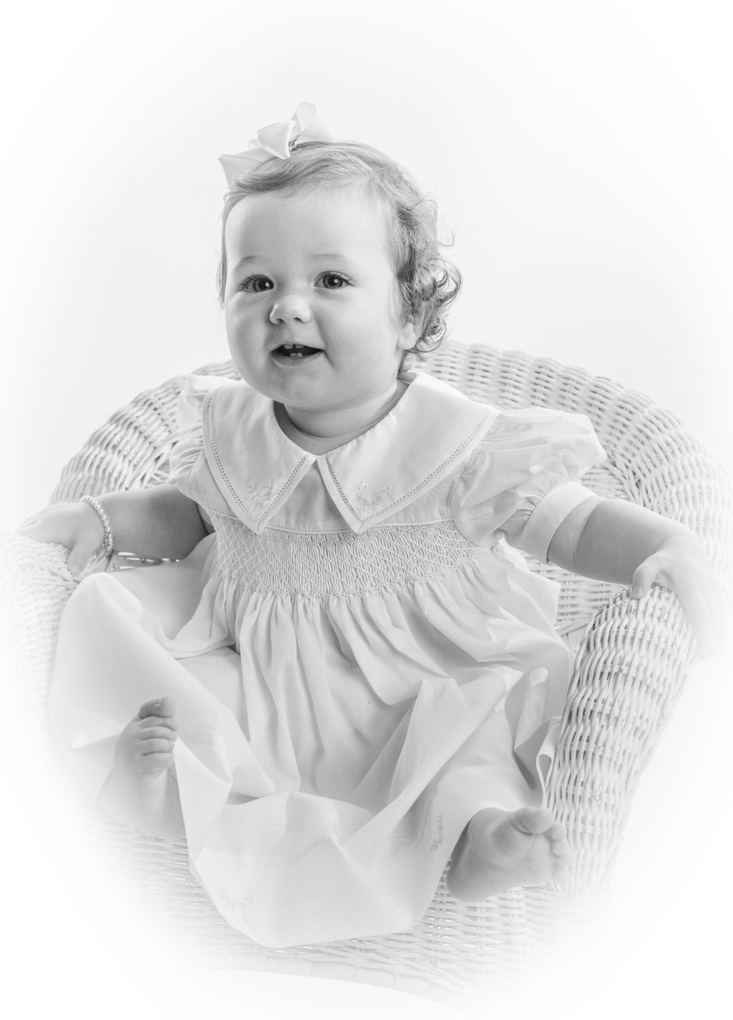 Black and white portrait of a smiling baby in a white smocked dress sitting in a wicker chair with a bow in her hair.