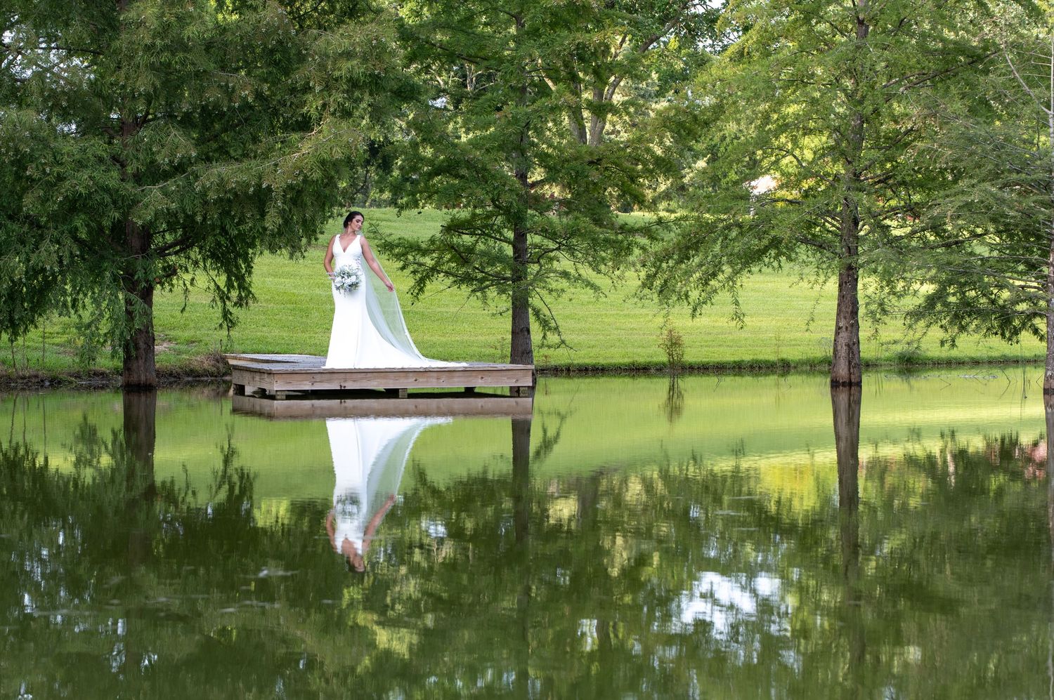 Bride in white gown standing on a dock reflected in a serene pond surrounded by lush green trees.