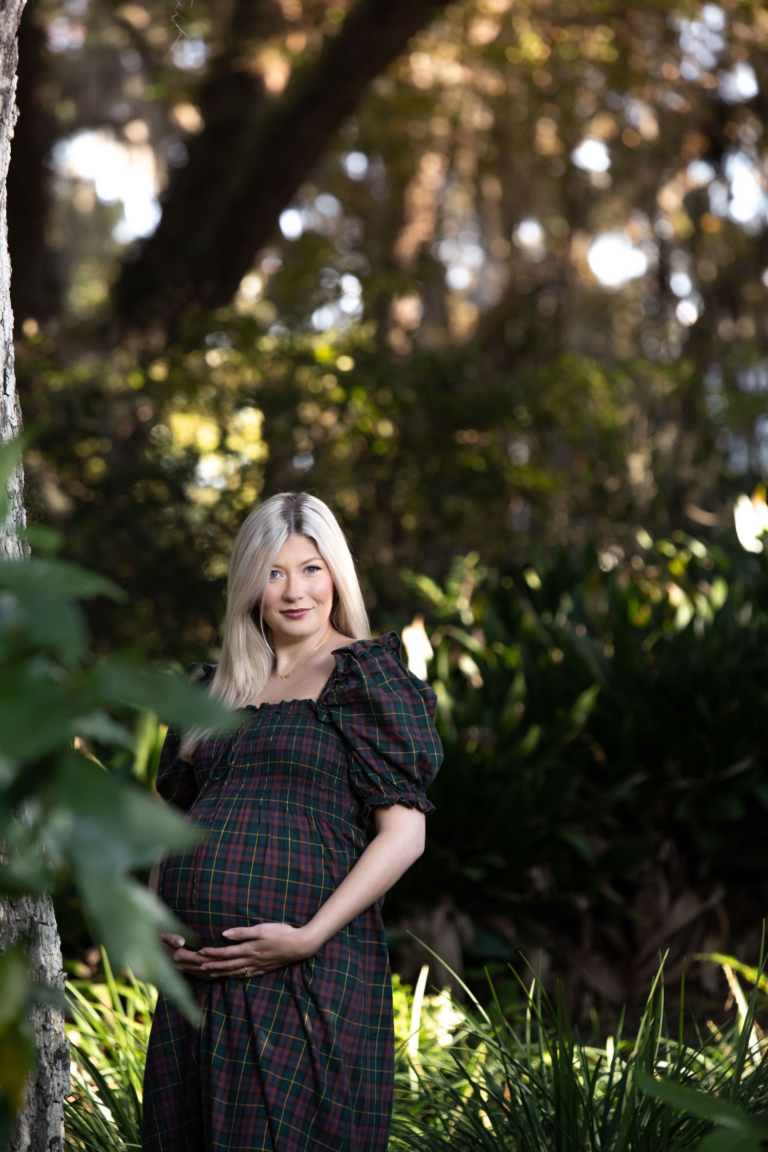 Pregnant woman in plaid dress cradling baby bump outdoors among lush green trees and foliage.