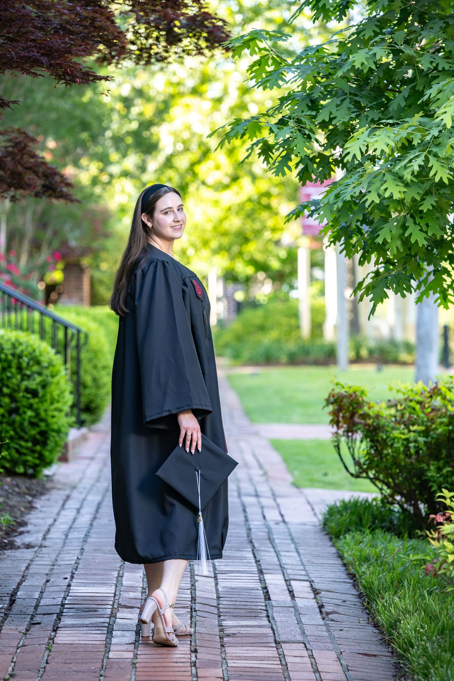 Young woman in black graduation gown and cap smiling on a brick pathway surrounded by lush green trees.