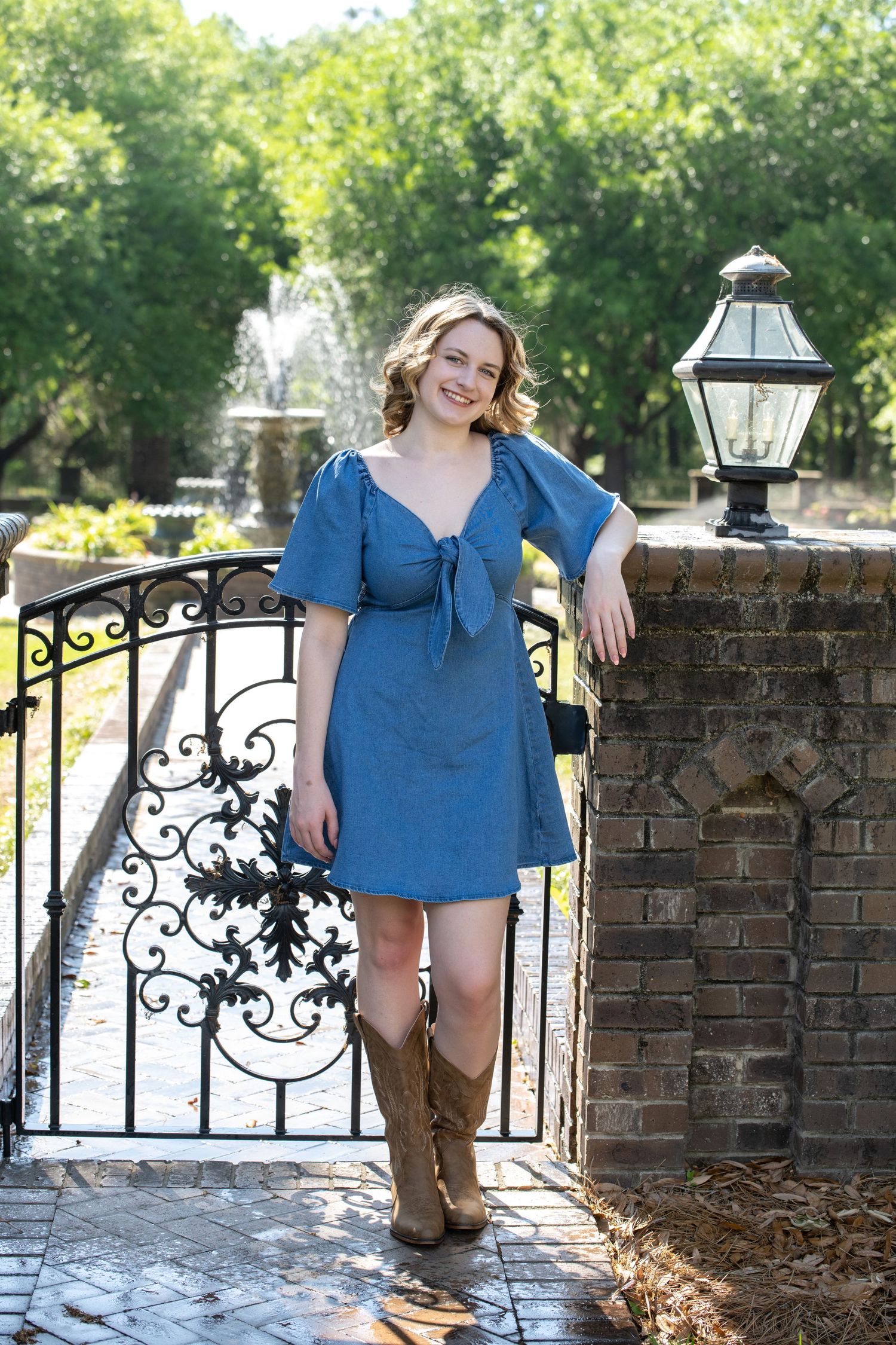 Young woman in blue dress and cowboy boots posing by ornate iron gate with brick pillar and fountain.